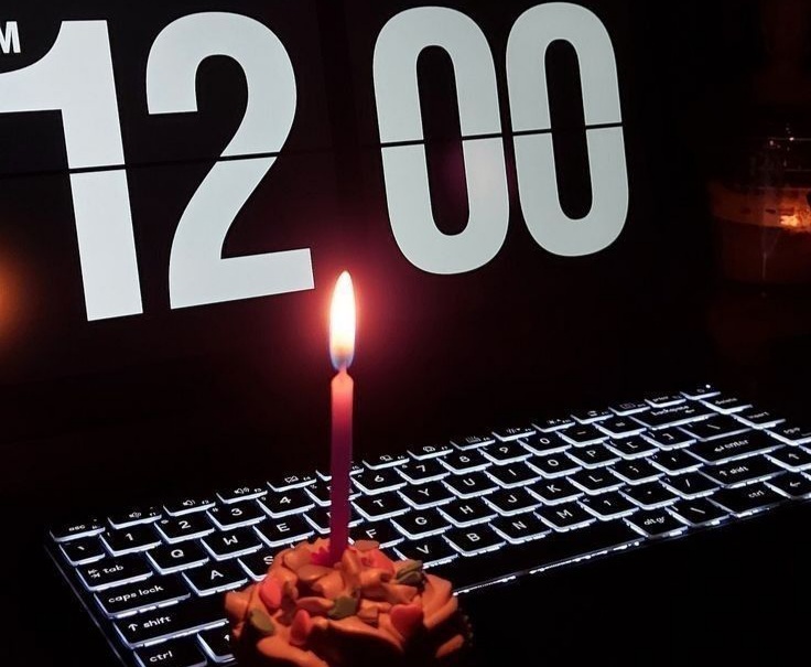 Foto sebuah lilin menyala di atas kue kecil dengan latar belakang layar monitor yang menampilkan angka jam digital 12:00, melambangkan momen pergantian tahun sebagai pengingat waktu yang terus berjalan
