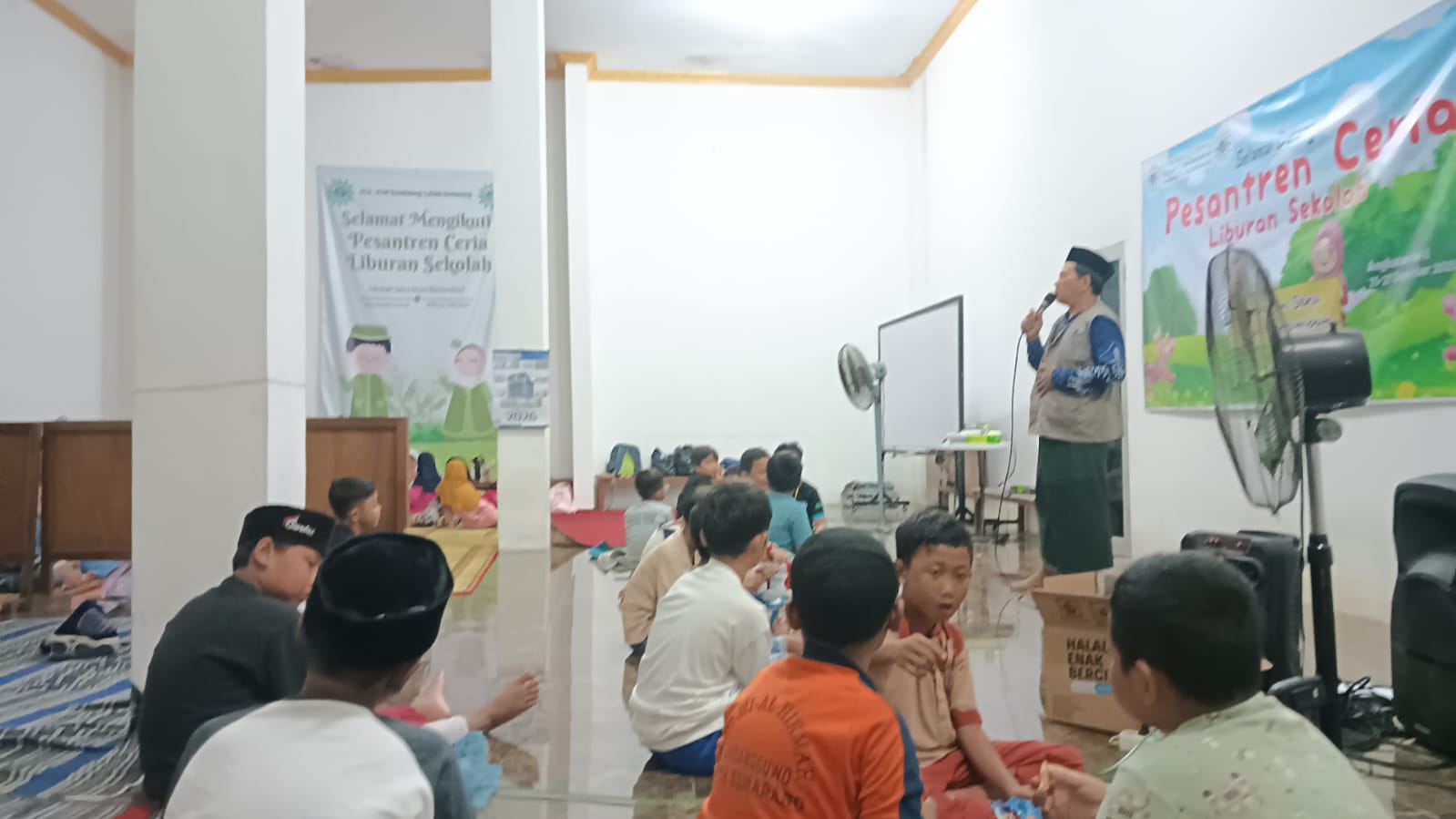 Suasana dalam ruangan Pesantren Ceria di Griya Harmoni Semarang, memperlihatkan seorang pemateri pria sedang berbicara di depan puluhan anak-anak yang duduk menyimak dengan latar belakang spanduk acara.