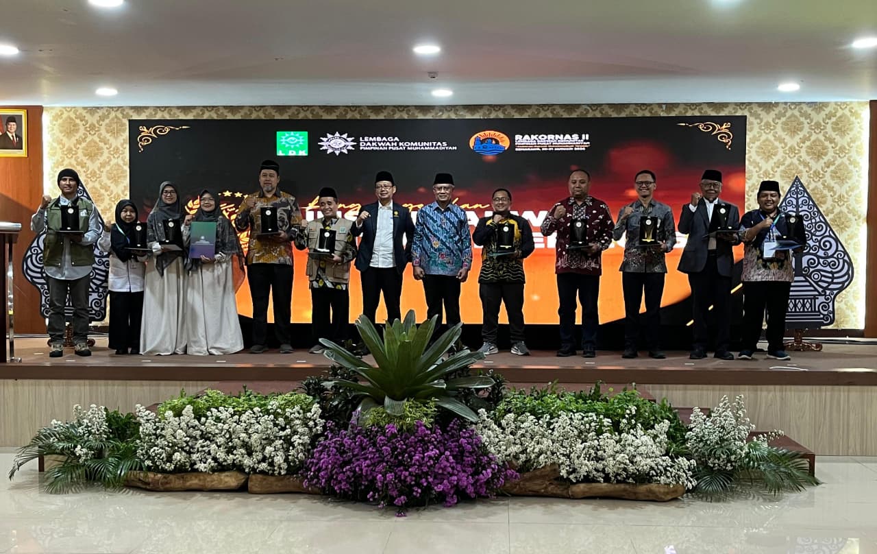 Sekelompok pria dan wanita berdiri berjajar di atas panggung dalam acara formal. Mereka memegang plakat penghargaan berwarna hitam dan emas. Di latar belakang terdapat layar digital besar bertuliskan 'Rakornas II Lembaga Dakwah Komunitas Pimpinan Pusat Muhammadiyah' dengan logo LDK dan Muhammadiyah.