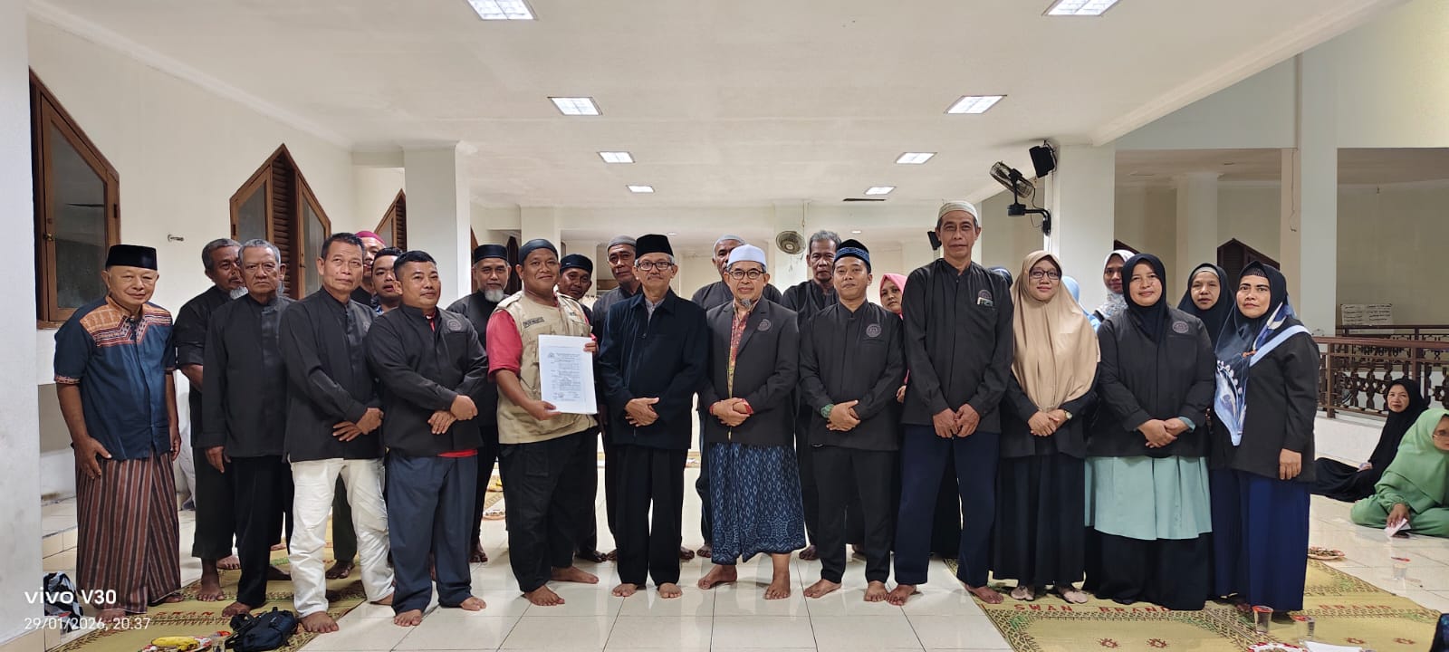 Foto bersama di dalam aula masjid yang luas dan bersih. Sekelompok pria dan wanita berjajar rapi mengenakan pakaian serba hitam dan batik. Di bagian tengah, seorang pria mengenakan rompi krem LDK sedang memegang map putih berisi dokumen pengukuhan relawan, diapit oleh tokoh takmir masjid. Suasana ruangan terang dengan plafon minimalis dan lampu kotak-kotak.