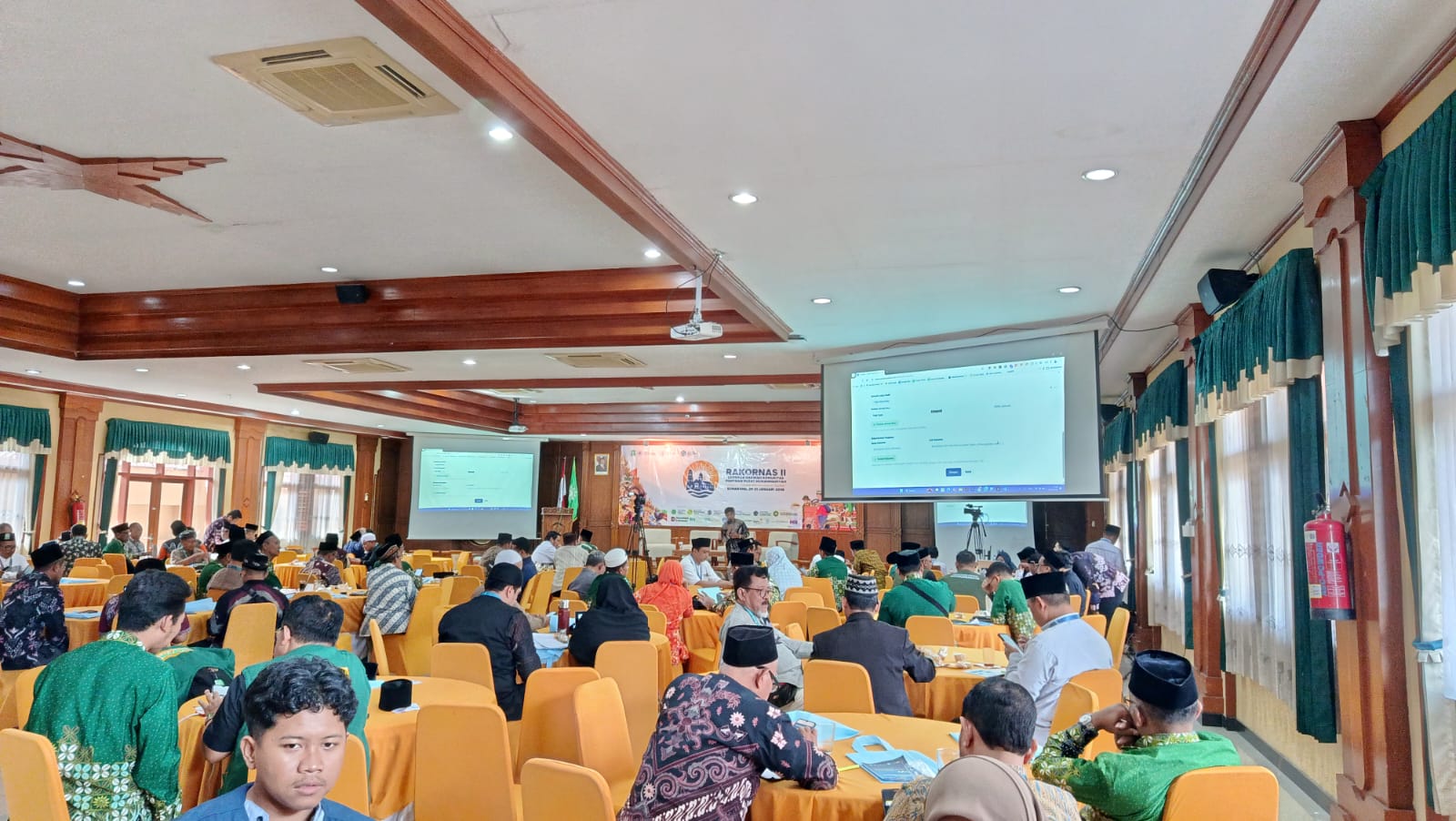 Suasana di dalam aula pertemuan yang luas dengan meja-meja bundar tertutup kain jingga. Para peserta pria dan wanita yang mengenakan batik Muhammadiyah hijau dan seragam formal duduk menyimak presentasi. Di depan ruangan, terdapat layar proyektor besar yang menampilkan antarmuka website SIDAKMU yang sedang dijelaskan oleh pengembang di atas panggung.
