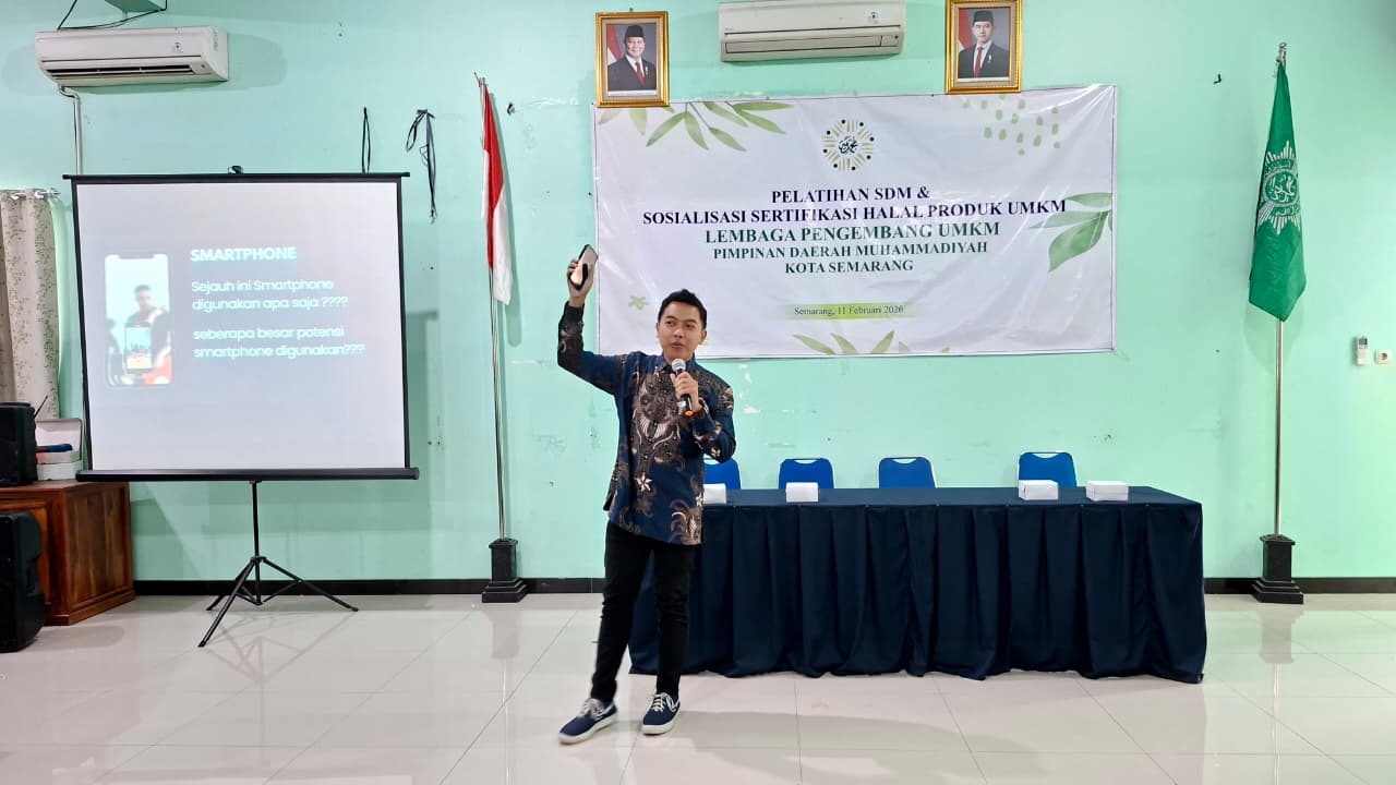Suasana acara pengajian dan sosialisasi di sebuah ruangan besar. Di latar belakang terdapat spanduk putih besar bertuliskan 'Pengajian KOMUNITAS Semarang, 01 Februari 2026' dengan foto Dr. H. AM Juma'i, SE., MM dan Dr. H. Iskandar Chang, SE. M. Si. Di tengah panggung, seorang pria mengenakan jas hitam, kemeja putih, peci hitam, dan sarung batik duduk di kursi kayu ukir besar sambil berbicara memegang mikrofon. Di lantai depan terdapat karpet merah bermotif bunga dan karpet hijau, serta beberapa jamaah pria duduk bersila di sisi kanan.