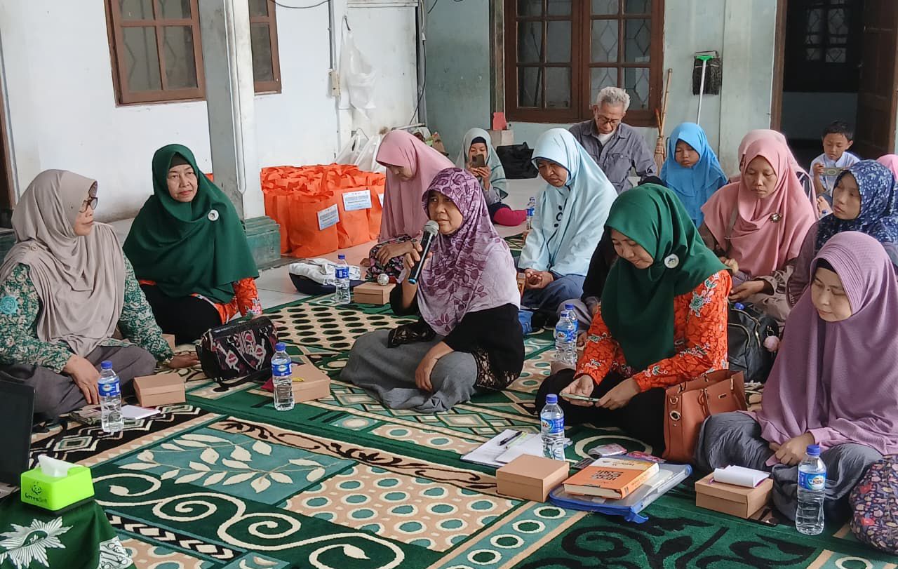 Sekelompok perempuan dan pria duduk bersila di atas karpet hijau bermotif di sebuah teras rumah. Seorang perempuan di tengah mengenakan kerudung bermotif bunga ungu sedang berbicara memegang mikrofon. Di sekelilingnya, para peserta perempuan mengenakan kerudung dengan berbagai warna seperti hijau, merah muda, dan biru sedang menyimak. Di latar belakang terlihat tumpukan tas belanja berwarna oranye (sembako) dan beberapa pria yang duduk menyimak. Suasana terasa khidmat dan kekeluargaan