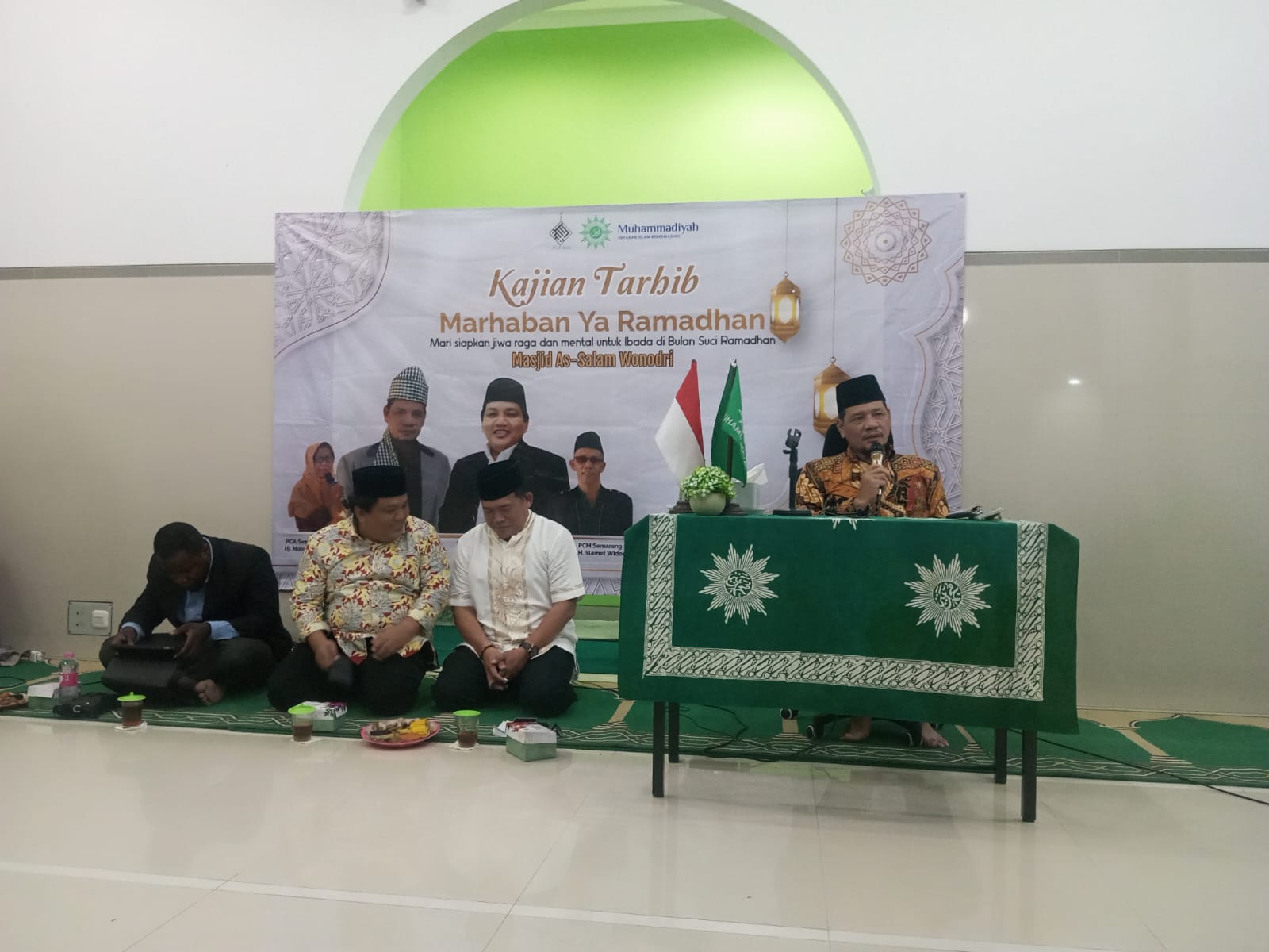 Suasana pengajian di dalam masjid dengan dinding putih dan aksen hijau pada lengkungan mihrab. Di tengah terdapat spanduk besar bertuliskan 'Kajian Tarhib Marhaban Ya Ramadhan' dengan foto beberapa tokoh. Seorang pria mengenakan batik cokelat dan peci hitam duduk di depan meja yang ditutup kain hijau berlogo Muhammadiyah, sedang berbicara menggunakan mikrofon. Di sampingnya, tiga orang pria duduk bersila di atas karpet hijau menyimak penjelasan. Di depan mereka tersedia beberapa kotak snack dan air mineral.