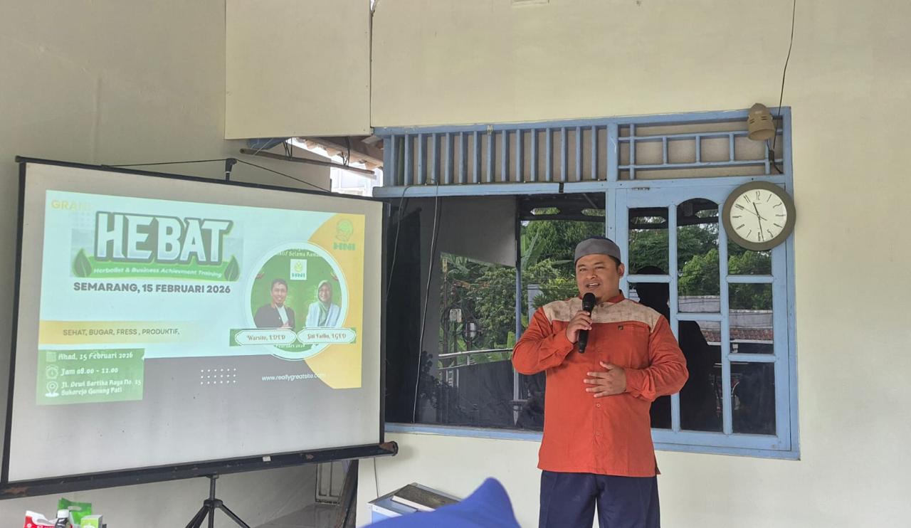 Seorang pria (Imron Hamzah) mengenakan kemeja oranye kecokelatan dengan aksen garis di bahu dan peci abu-abu, berdiri sambil memegang mikrofon dengan tangan kanan dan tangan kiri diletakkan di perut. Ia berdiri di samping layar proyektor putih yang menampilkan slide berjudul 'HEBAT: Herbalist & Business Achievement Training' dengan latar hijau kekuningan, tanggal 15 Februari 2026, dan foto narasumber Warsito serta Siti Fadilah. Latar belakang berupa dinding putih dengan jendela kayu biru dan jam dinding yang menunjukkan pukul 11.55. Suasana terlihat seperti ruang pertemuan semi-terbuka.