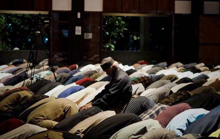 Foto dari arah belakang penceramah yang mengenakan batik cokelat dan peci hitam di atas podium kayu, sedang berbicara di hadapan ratusan jamaah pria yang duduk bersila di karpet merah dalam aula masjid yang luas.