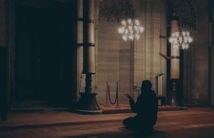 Foto siluet seorang pria yang sedang duduk bersimpuh dan berdoa dengan mengangkat kedua tangan di dalam sebuah masjid yang luas dan gelap. Pencahayaan hangat berasal dari dua buah lampu gantung hias yang estetik, menciptakan suasana yang syahdu dan kontemplatif.