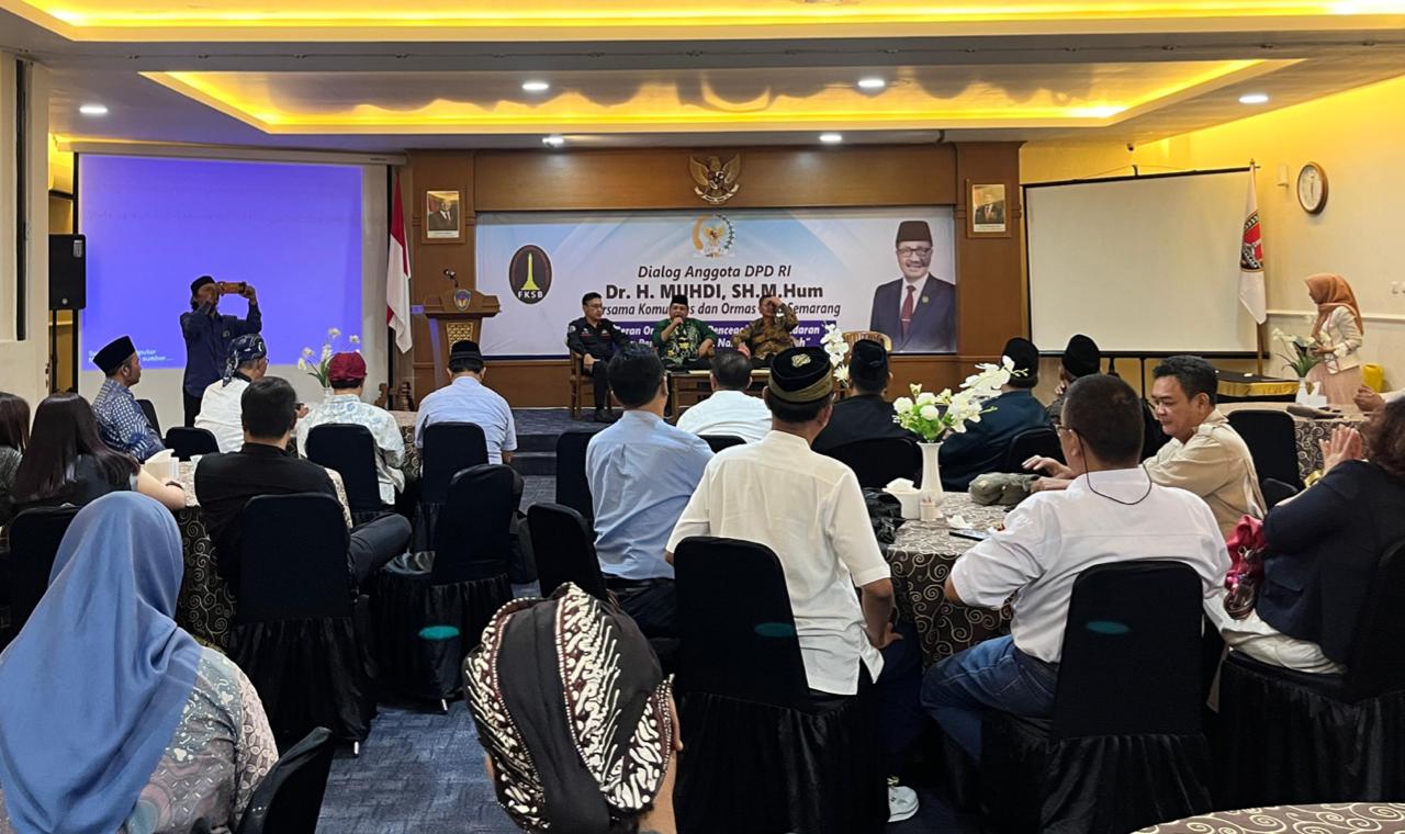 Foto suasana dialog publik di dalam sebuah aula hotel dengan pencahayaan hangat. Tampak dari arah belakang, puluhan peserta mengenakan kemeja dan pakaian batik duduk di kursi-kursi yang mengelilingi meja bundar dengan taplak motif cokelat. Di depan, terdapat panggung dengan spanduk besar bertuliskan "Dialog Anggota DPD RI Dr. H. MUHDI, SH., M.Hum. bersama Komunitas dan Ormas Semarang".