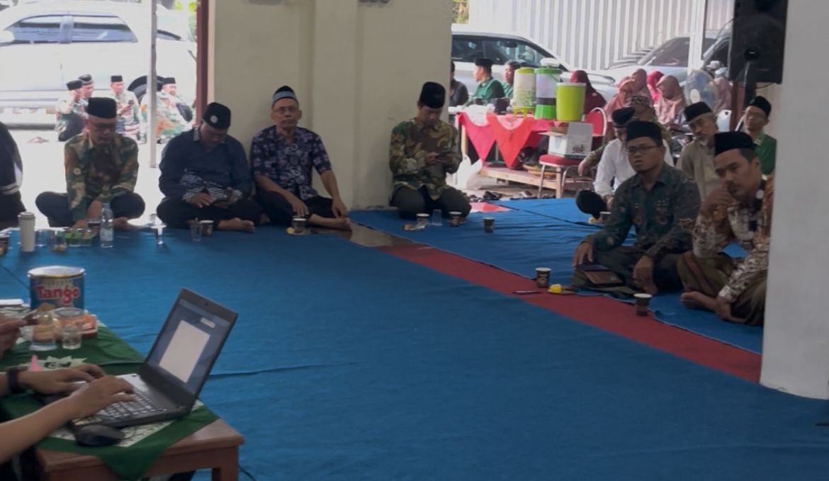 Foto suasana rapat koordinasi di dalam ruangan beralaskan karpet biru (lesehan). Terlihat sejumlah pria mengenakan peci hitam dan kemeja batik atau koko sedang duduk melingkar. Di sudut kiri bawah, tampak tangan seseorang sedang mengoperasikan laptop di atas meja kecil berbajukan taplak hijau dengan logo matahari Muhammadiyah. Di tengah ruangan, terdapat beberapa gelas minuman plastik dan kaleng biskuit. Di latar belakang, terlihat pintu terbuka yang memperlihatkan area parkir dengan beberapa mobil putih dan jemaah lain yang sedang berbincang di luar ruangan.