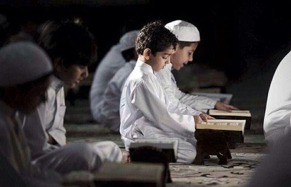 Foto suasana syahdu di dalam masjid pada malam hari dengan pencahayaan minim yang dramatis (low light). Tampak sekelompok anak laki-laki mengenakan baju koko putih dan peci putih sedang duduk bersila di atas karpet motif. Fokus utama tertuju pada seorang anak di tengah yang sedang menunduk khusyuk membaca mushaf Al-Quran di atas meja kayu kecil (rehal).