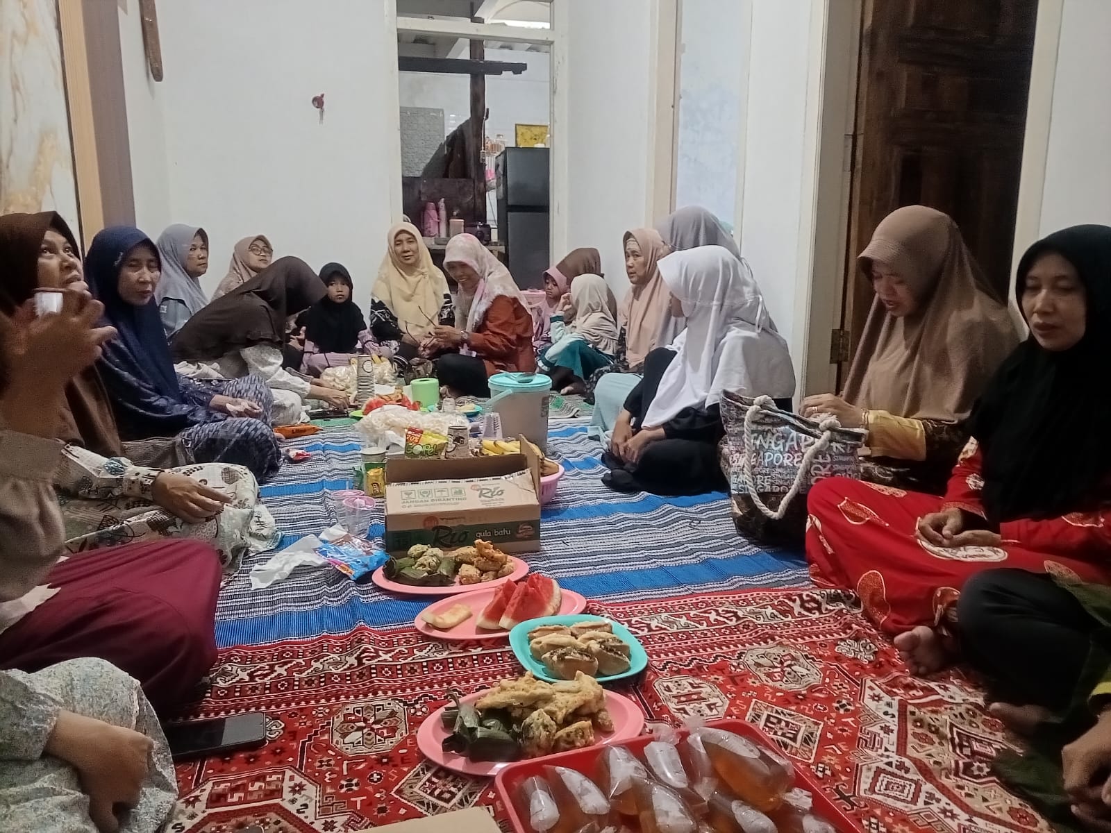 Foto sekelompok wanita (guru dan keluarga) yang mengenakan jilbab duduk melingkar di atas karpet bermotif merah dan biru di dalam sebuah ruangan rumah. Di tengah-tengah mereka tersaji aneka hidangan takjil seperti gorengan, irisan semangka, kue basah, dan minuman plastik yang diletakkan di piring-piring plastik. Suasana tampak akrab dan santai saat mereka berbincang menunggu waktu berbuka. Di latar belakang terlihat pintu kayu dan area dapur rumah.