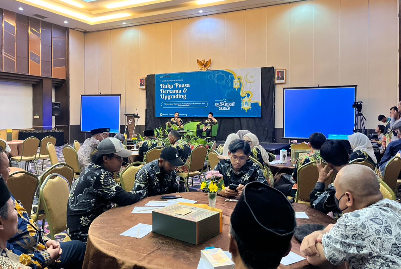 Foto suasana di dalam Ballroom Hotel Grasia Semarang yang luas. Di depan ruangan terdapat panggung dengan spanduk biru besar bertuliskan "Buka Puasa Bersama & Upgrading" serta logo PT Sinar Muhindo Konstruksi dan tanggal 13 Maret 2026. Di atas panggung, dua orang pria mengenakan seragam batik hitam bermotif sedang duduk di kursi hijau sambil berbincang. Di depan panggung, para peserta pria dan wanita yang mengenakan seragam batik serupa duduk melingkar mengelilingi meja-meja bundar yang ditutupi kain cokelat. Terdapat dua layar biru besar di sisi kiri dan kanan panggung.