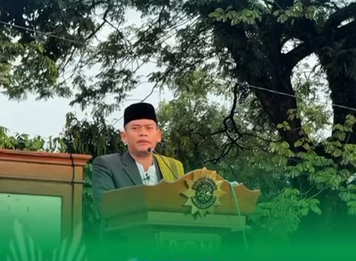 Foto close-up dari arah bawah yang memperlihatkan seorang pria Muslim mengenakan peci hitam, kemeja putih, jas abu-abu tua, dan syal batik kuning keemasan yang diselempangkan di bahu kirinya. Ia sedang berdiri tegak di atas podium kayu berukir yang menampilkan logo matahari putih Muhammadiyah dengan latar belakang daun pohon beringin besar yang rimbun dan langit pagi yang cerah. Wajahnya menatap ke depan dengan ekspresi serius saat memberikan khotbah, dan terdapat mikrofon kecil yang terpasang di kerah kemejanya.