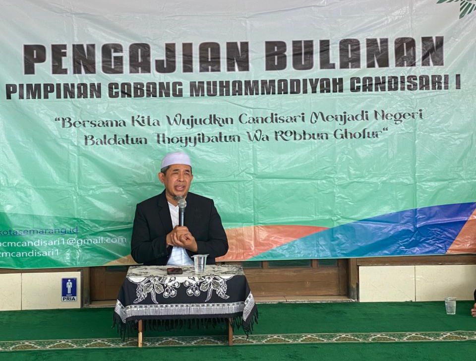 Foto memperlihatkan KH Drs. Muh Saidun, M.Ag. sedang memberikan ceramah di depan sebuah backdrop besar berwarna hijau dan putih. Beliau mengenakan jas hitam, kemeja putih, dan peci putih (haji), sedang memegang mikrofon dengan kedua tangannya. Di belakang beliau, backdrop bertuliskan "PENGAJIAN BULANAN PIMPINAN CABANG MUHAMMADIYAH CANDISARI 1" dengan kutipan tema "Bersama Kita Wujudkan Candisari Menjadi Negeri Baldatun Thoyyibatun Wa Robbun Ghofur". Beliau duduk di balik meja kecil yang dilapisi taplak batik bermotif parang, dengan sebuah gelas air mineral di atasnya. Lantai ruangan dilapisi karpet hijau bermotif sajadah. Di sudut kiri bawah backdrop, terlihat informasi kontak digital organisasi.