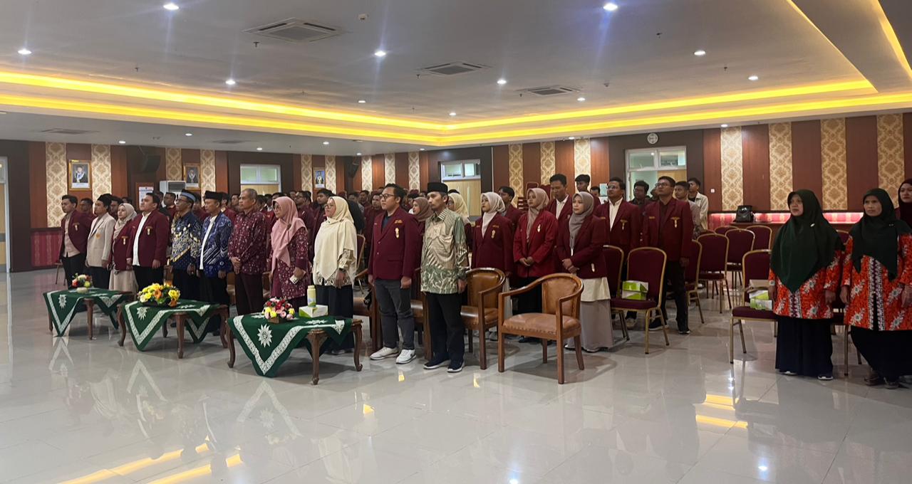 Suasana formal di dalam Aula Lantai 8 GKB 2 Unimus yang luas dengan pencahayaan hangat dari lampu plafon. Puluhan peserta pria dan wanita berdiri rapi mengikuti prosesi pembukaan, sebagian besar mengenakan jas almamater IMM berwarna merah marun. Di barisan depan, tampak sejumlah tokoh pimpinan mengenakan pakaian formal seperti batik dan kemeja. Ruangan didekorasi dengan panel dinding bermotif geometris dan terdapat meja-meja kecil dengan hiasan bunga di bagian depan. Atmosfer foto menunjukkan kekhidmatan acara skala nasional.