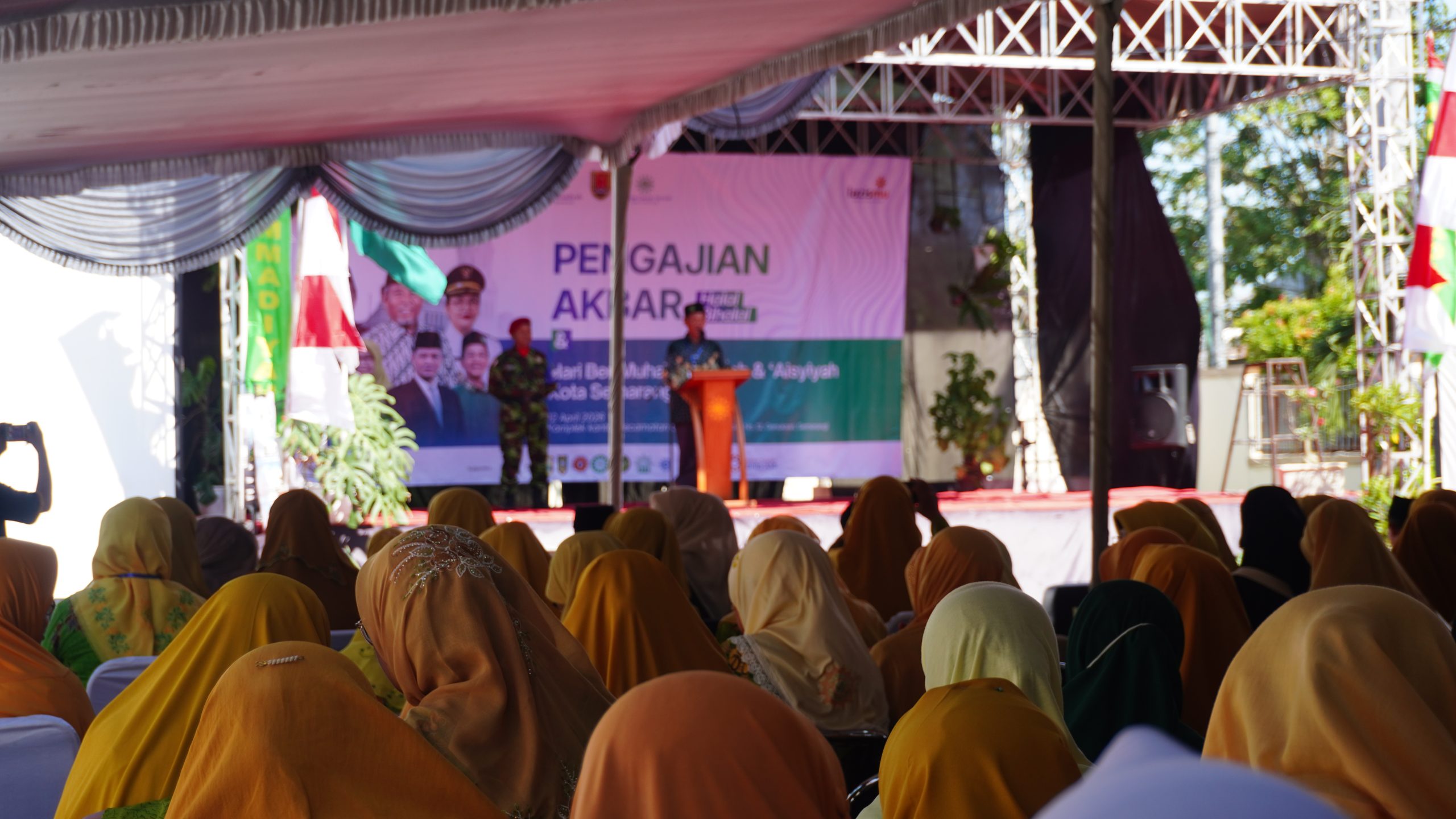 Foto suasana pengajian akbar dari sudut pandang belakang jamaah yang didominasi oleh wanita mengenakan jilbab berwarna kuning mustard dan hijau. Para jamaah duduk rapi di bawah tenda putih besar yang menghadap ke arah panggung utama. Di atas panggung, seorang pria sedang berbicara di podium, didampingi oleh personel keamanan berseragam loreng yang berjaga di sisi kiri panggung. Latar belakang panggung menampilkan spanduk besar bertuliskan "PENGAJIAN AKBAR" dengan logo Muhammadiyah dan 'Aisyiyah. Cahaya matahari yang cerah masuk dari sisi kanan tenda, menciptakan suasana acara luar ruangan yang tertib.