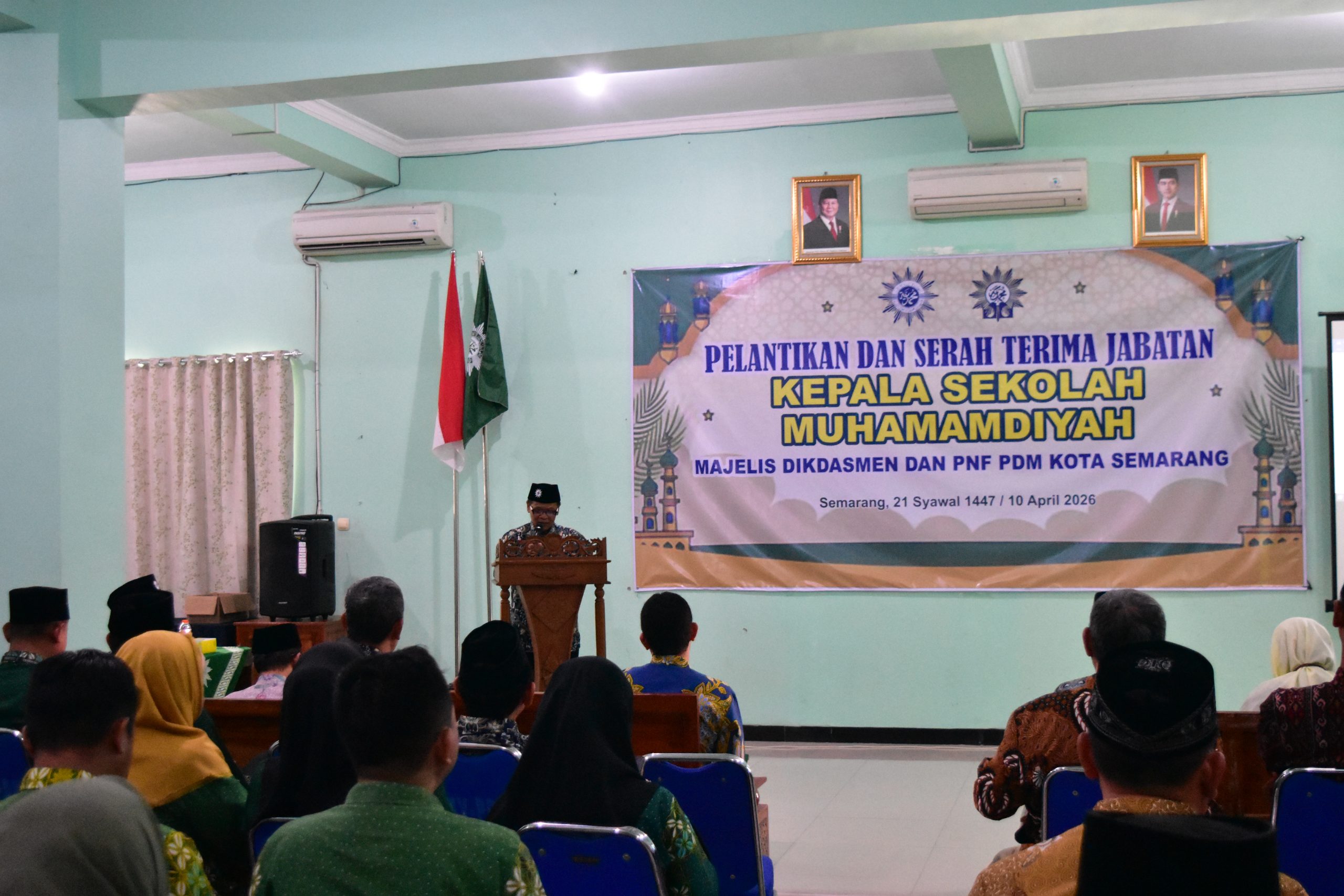 Foto suasana acara resmi di dalam sebuah ruangan pertemuan dengan dinding berwarna hijau muda. Di bagian depan, terdapat spanduk putih besar dengan tulisan biru dan kuning: "PELANTIKAN DAN SERAH TERIMA JABATAN KEPALA SEKOLAH MUHAMMADIYAH MAJELIS DIKDASMEN DAN PNF PDM KOTA SEMARANG" bertanggal 10 April 2026. Di bawah spanduk, seorang pria berpeci hitam berdiri di balik podium kayu sedang memberikan sambutan. Di samping kiri podium, terdapat bendera Merah Putih dan bendera hijau Muhammadiyah. Di bagian atas spanduk, terpasang foto resmi Presiden dan Wakil Presiden RI serta sebuah unit AC. Di latar depan, tampak deretan tamu undangan yang duduk di kursi biru sedang menyimak acara dengan khidmat.