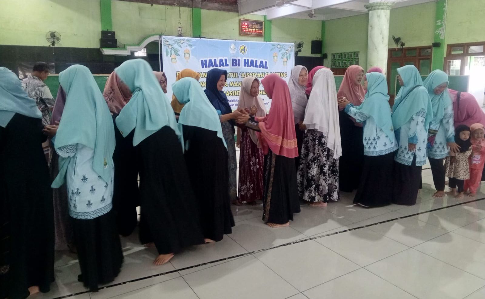 Foto suasana hangat di dalam Masjid Al Muhajirin Dondong yang menunjukkan puluhan kader perempuan Nasyiatul Aisyiyah sedang melakukan tradisi mushofahah (bersalam-salaman). Mereka berbaris memanjang membentuk antrean yang tertib, mengenakan jilbab seragam berwarna biru muda dan hijau toska, serta pakaian muslimah bermotif batik. Di latar belakang, tampak sebuah banner biru bertuliskan "HALAL BI HALAL" dengan logo Nasyiatul Aisyiyah dan Aisyiyah yang terpasang di dinding masjid berwarna hijau muda. Ruangan masjid terlihat bersih dengan lantai keramik putih dan pilar besar di sisi kanan. Di ujung antrean, terlihat beberapa anak kecil ikut mendampingi ibunya dalam suasana yang penuh keakraban.