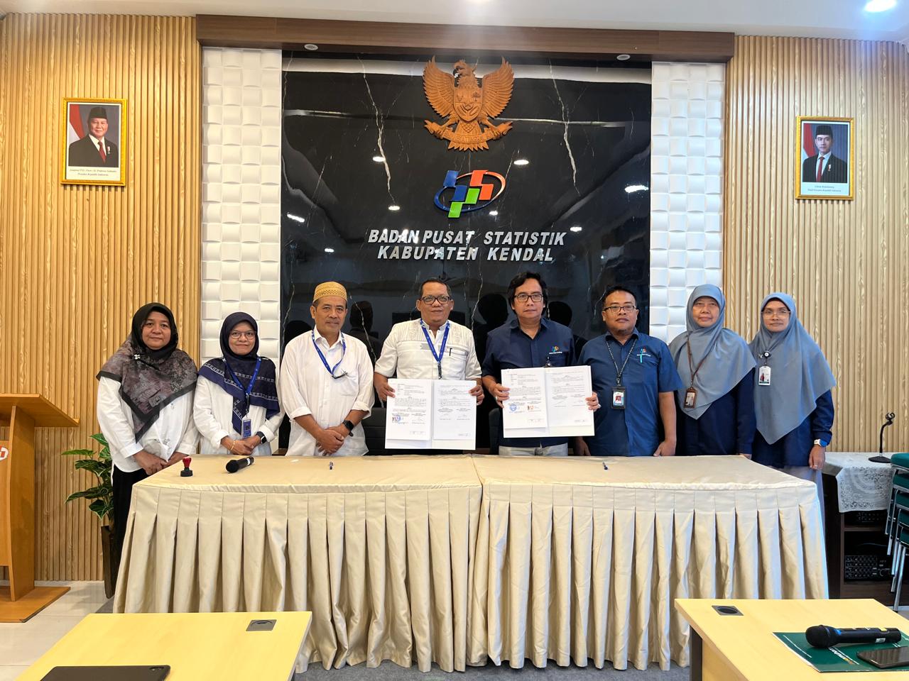 Foto bersama delapan orang di dalam kantor Badan Pusat Statistik (BPS) Kabupaten Kendal. Di latar belakang, terdapat dinding kayu dengan logo BPS, lambang Garuda Pancasila, serta foto resmi Presiden dan Wakil Presiden RI. Dua pria di tengah tampak memegang dokumen resmi sebagai simbol kerja sama. Peserta yang hadir terdiri dari pria dan wanita, beberapa mengenakan seragam biru BPS dan lainnya mengenakan pakaian formal bebas. Suasana ruangan tampak formal dengan meja panjang yang ditutupi kain krem di bagian depan.