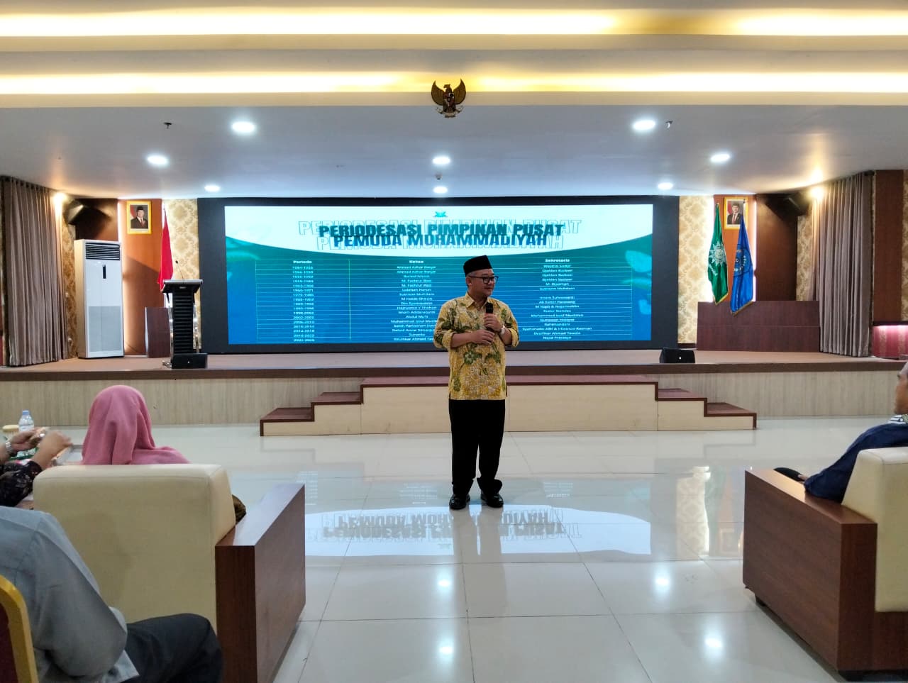 Foto suasana di dalam aula besar (Aula GKB 2 Unimus) dengan pencahayaan terang dari lampu plafon yang memanjang. Di tengah panggung, berdiri Prof. Dr. KH. Abdul Mu'ti, M.Ed., mengenakan kemeja batik bermotif warna kuning dan hijau serta peci hitam, sedang memegang mikrofon dan memberikan sambutan. Di belakang beliau, terdapat layar LED raksasa yang menampilkan slide presentasi berjudul "PERIODESASI PIMPINAN PUSAT PEMUDA MUHAMMADIYAH" dengan latar belakang hijau dan biru. Di sisi kiri panggung terdapat bendera Merah Putih, dan di sisi kanan terdapat bendera Muhammadiyah serta bendera IMM. Beberapa peserta terlihat duduk di sofa krem di bagian depan, menyaksikan acara dengan khidmat.