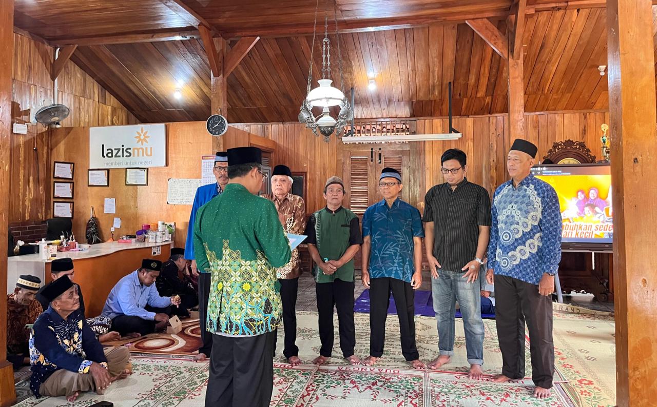 Foto suasana pelantikan di dalam sebuah ruangan berarsitektur kayu tradisional (joglo) yang hangat. Enam orang pria berdiri berjajar mengenakan peci hitam dan kemeja batik atau koko dengan berbagai motif. Di hadapan mereka, seorang pria berpunggung kamera yang mengenakan kemeja batik hijau motif parang sedang membacakan naskah pelantikan. Di latar belakang, terlihat logo "lazismu" berwarna oranye di dinding kayu, sebuah jam dinding menunjukkan pukul 06.00 pagi, dan beberapa jemaah lain duduk lesehan di atas karpet bermotif bunga. Sebuah layar televisi di sudut kanan menampilkan slide presentasi berwarna kuning.