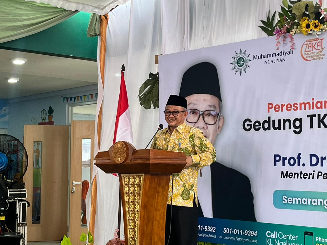 Foto memperlihatkan Menteri Pendidikan Dasar dan Menengah (Mendikdasmen) RI, Prof. Dr. KH. Abdul Mu'ti, M.Ed., sedang berdiri memberikan sambutan di balik podium kayu cokelat yang memiliki ukiran logo matahari Muhammadiyah di bagian depan. Beliau mengenakan kemeja batik bermotif parang dominan warna kuning dan hijau serta peci hitam. Di latar belakang panggung, terdapat banner putih besar bertuliskan "Peresmian Gedung TK" dengan foto wajah Prof. Abdul Mu'ti yang besar. Di sisi kiri panggung tampak bendera Merah Putih, dan di bagian bawah banner terlihat beberapa nomor kontak serta tulisan "LAZISMU". Suasana panggung dihiasi dekorasi kain putih dan rangkaian bunga di sudut kanan atas.