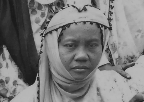 Potret close-up hitam putih Siti Munjiyah, tokoh perempuan Muhammadiyah, menatap lurus ke kamera. Ia mengenakan penutup kepala berwarna terang yang melilit di leher dan wajah, dengan hiasan manik-manik gelap di sepanjang pinggirannya. Latar belakang menunjukkan bagian dari pakaian bermotif bunga dan sebagian tangan orang lain di sisi kanan.