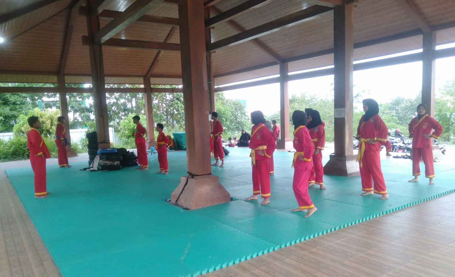 Foto sekelompok remaja pria dan wanita mengenakan seragam bela diri Tapak Suci berwarna merah khas dengan aksen kuning. Mereka sedang menjalani sesi latihan di dalam sebuah pendopo terbuka yang luas dengan lantai beralaskan matras puzzle berwarna hijau. Para siswa tampak berdiri dalam barisan yang teratur, melakukan gerakan pemanasan atau kuda-kuda di bawah naungan struktur bangunan kayu yang kokoh. Di latar belakang terlihat pemandangan pepohonan hijau yang asri dan beberapa sepeda motor yang terparkir