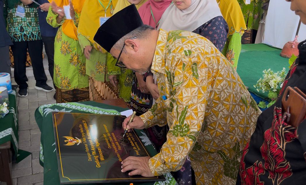 Foto close-up sudut tinggi yang memperlihatkan Menteri Pendidikan Dasar sekaligus Sekum PP Muhammadiyah, Prof. Dr. KH. Abdul Mu'ti, M.Ed., sedang membungkuk untuk menandatangani prasasti peresmian. Beliau mengenakan kemeja batik bermotif parang dominan warna kuning dan hijau, serta peci hitam. Tangan kanannya memegang pena emas di atas prasasti hitam dengan tulisan berwarna emas yang berbunyi "Gedung TK Aisyiyah Bustanul Athfal 31 Wonosari Ngaliyan". Di latar belakang, tampak barisan ibu-ibu pengurus Aisyiyah mengenakan seragam batik hijau dan jilbab kuning cerah sedang menyaksikan prosesi tersebut dengan khidmat.