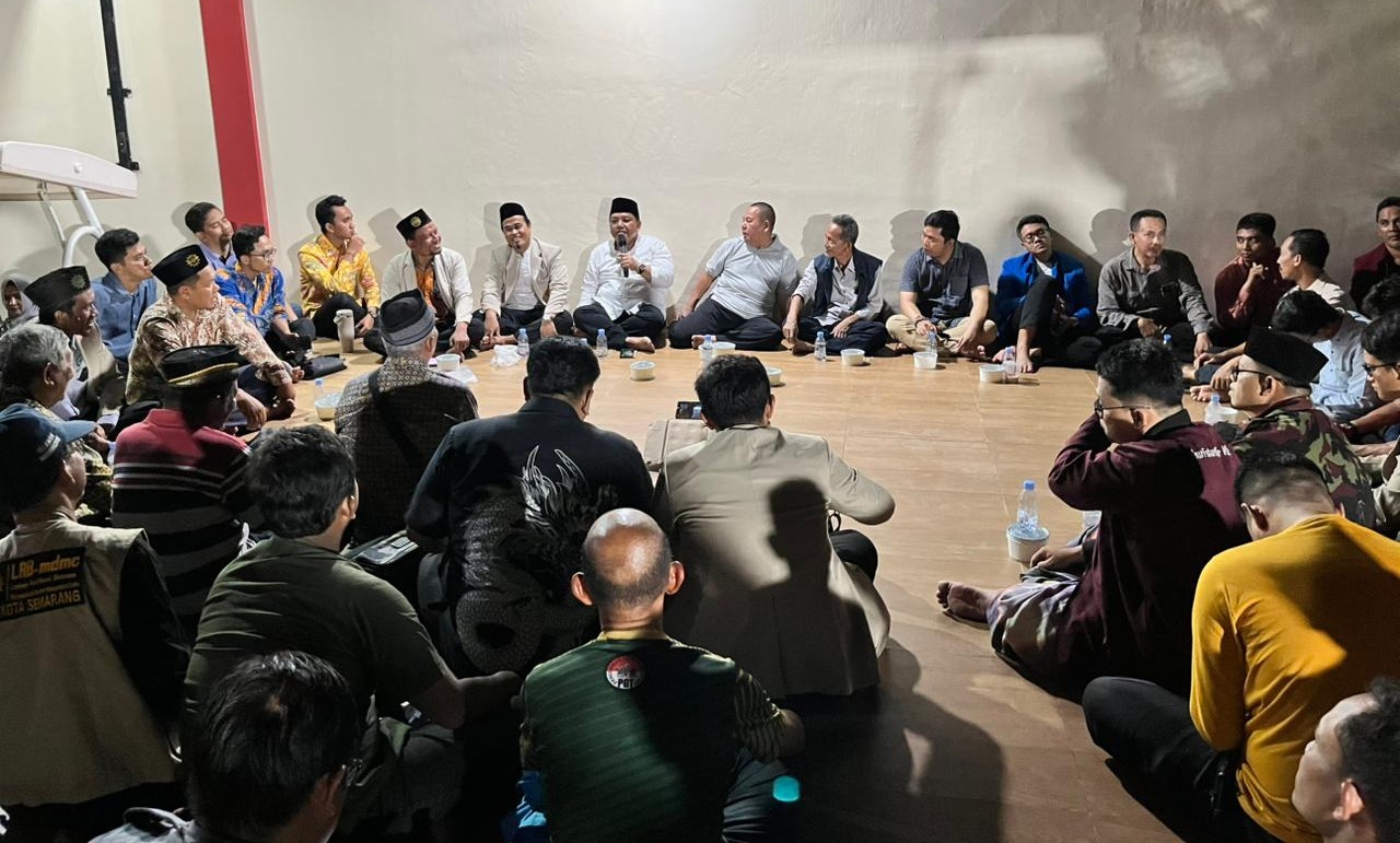Foto suasana pertemuan formal namun santai yang dilakukan dengan duduk bersila (lesehan) membentuk lingkaran besar di sebuah ruangan. Di bagian tengah latar belakang, seorang pria berbaju putih dan peci hitam sedang berbicara memegang mikrofon, diapit oleh para tokoh senior lainnya. Peserta yang hadir didominasi oleh pria yang mengenakan pakaian beragam, mulai dari kemeja batik, baju koko, jas organisasi, hingga seragam relawan MDMC (Muhammadiyah Disaster Management Center). Ruangan memiliki pencahayaan temaram dengan dinding berwarna krem, menciptakan suasana diskusi yang hangat dan akrab pada malam hari. Di depan para peserta terdapat botol air mineral dan mangkuk makanan kecil.