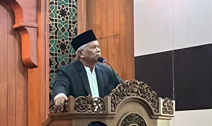 Foto close-up seorang pria paruh baya mengenakan jas hitam dan peci hitam sedang memberikan ceramah dari balik mimbar kayu yang dihiasi ukiran detail. Beliau berbicara di depan mikrofon dengan ekspresi serius namun tenang, menatap ke arah depan. Di latar belakang, terlihat bagian dinding masjid dengan motif geometris bintang delapan (khas dekorasi Islami) berwarna emas dan hijau yang memberikan kesan megah dan sakral. Pencahayaan fokus pada sosok penceramah, menciptakan suasana yang khusyuk.