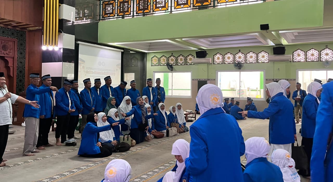 Foto suasana di dalam masjid yang luas dengan dominasi warna hijau muda dan pencahayaan terang. Puluhan calon jemaah haji, baik pria maupun wanita, terlihat mengenakan seragam batik berwarna biru cerah dengan motif khas. Sebagian jemaah wanita mengenakan jilbab putih, sementara jemaah pria mengenakan peci hitam. Di bagian depan, terdapat seorang pria yang sedang berbicara menggunakan mikrofon, memberikan arahan kepada para jemaah yang berdiri dan duduk bersila di atas karpet masjid. Layar proyektor di latar belakang menampilkan informasi mengenai keberangkatan haji. Ekspresi para jemaah menunjukkan antusiasme dan rasa haru.