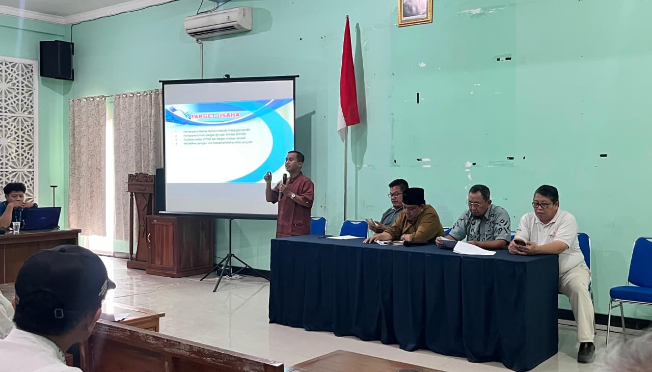 Foto suasana sosialisasi di dalam sebuah aula dengan dinding berwarna hijau muda. Di depan ruangan, seorang pria mengenakan kemeja merah marun sedang berdiri memberikan presentasi di samping layar proyektor yang menampilkan teks "TARGET USAHA". Di sebelah kanannya, terdapat empat orang pria duduk di balik meja panjang yang ditutupi kain biru tua, dengan bendera Merah Putih berdiri tegak di belakang mereka. Peserta rapat terlihat duduk di bangku kayu di bagian depan, menyimak paparan materi.