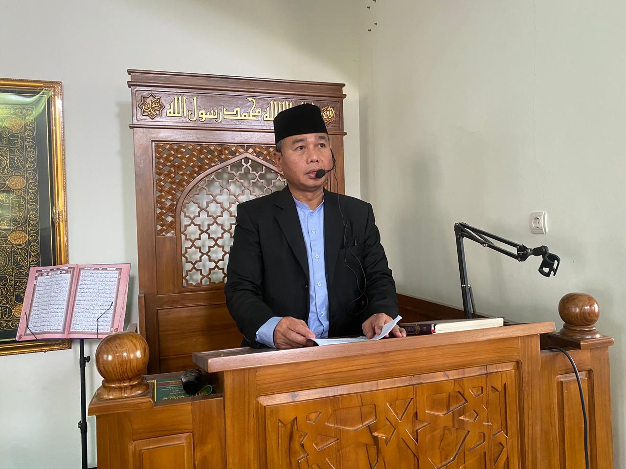 Foto seorang pria (Mukti Warso, M.M.) mengenakan jas hitam, kemeja biru muda, dan peci hitam sedang berdiri di atas mimbar kayu yang dihiasi ukiran khas Islami. Pria tersebut menggunakan mikrofon headset dan kedua tangannya memegang naskah di atas meja mimbar. Di sisi kiri mimbar, tampak sebuah papan informasi dengan teks Arab dan di dinding belakang terdapat saklar lampu serta kabel mikrofon yang menjuntai. Suasana foto menunjukkan momen penyampaian khutbah atau tausiyah di dalam ruangan masjid.