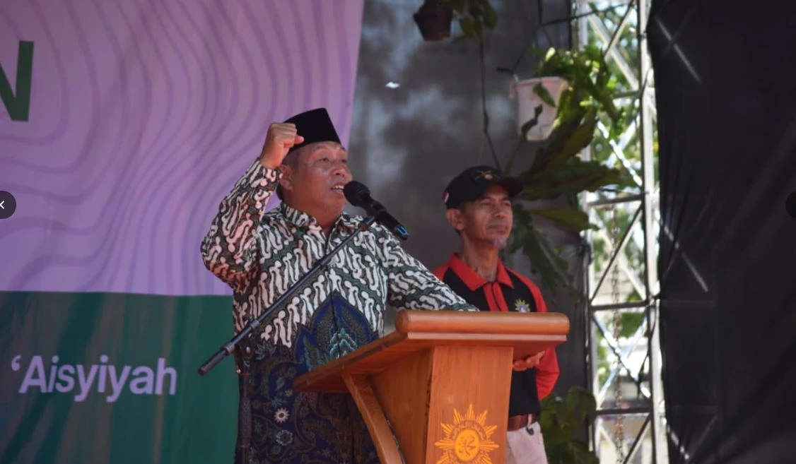 Dr. Agus Taufiqurrahman, Ketua PP Muhammadiyah, mengenakan kemeja batik dan peci hitam, mengangkat tangan kanannya yang mengepal kuat dalam pose orasi bersemangat di podium kayu berlogo Muhammadiyah. Di latar belakang kiri terdapat spanduk dengan teks 'Aisyiyah terlihat.