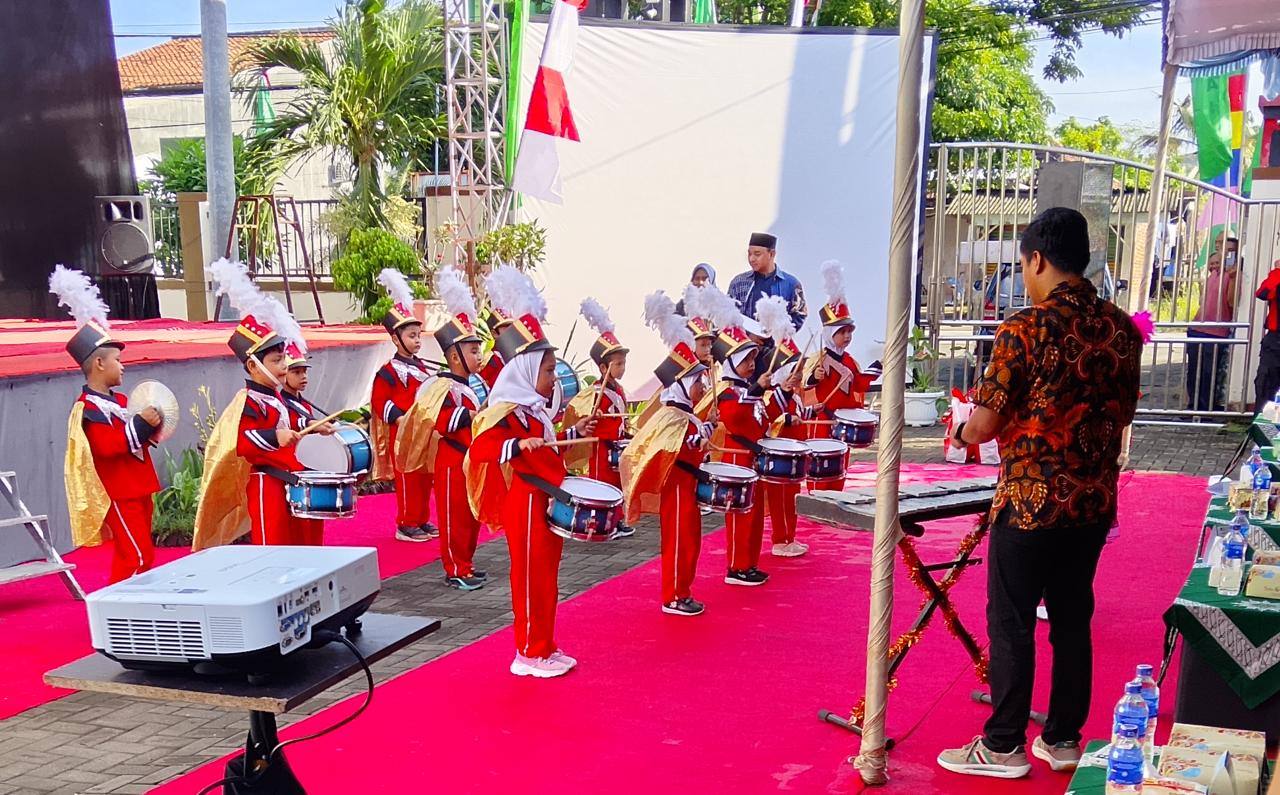 Foto luar ruangan yang memperlihatkan sekelompok anak kecil (siswa TK) mengenakan seragam drumband berwarna merah cerah dengan aksen emas dan topi tinggi berhias bulu putih. Mereka berdiri di atas karpet merah panjang di sebuah area terbuka. Beberapa anak tampak memegang drum kecil berwarna biru, sementara yang lain memegang simbal. Di sisi kanan, seorang pria dewasa berbaju batik cokelat berdiri membelakangi kamera, tampak sedang memberikan instruksi atau memandu penampilan mereka di hadapan meja tamu. Di latar belakang, terlihat pepohonan hijau, tiang bendera, dan layar putih besar di samping panggung utama.