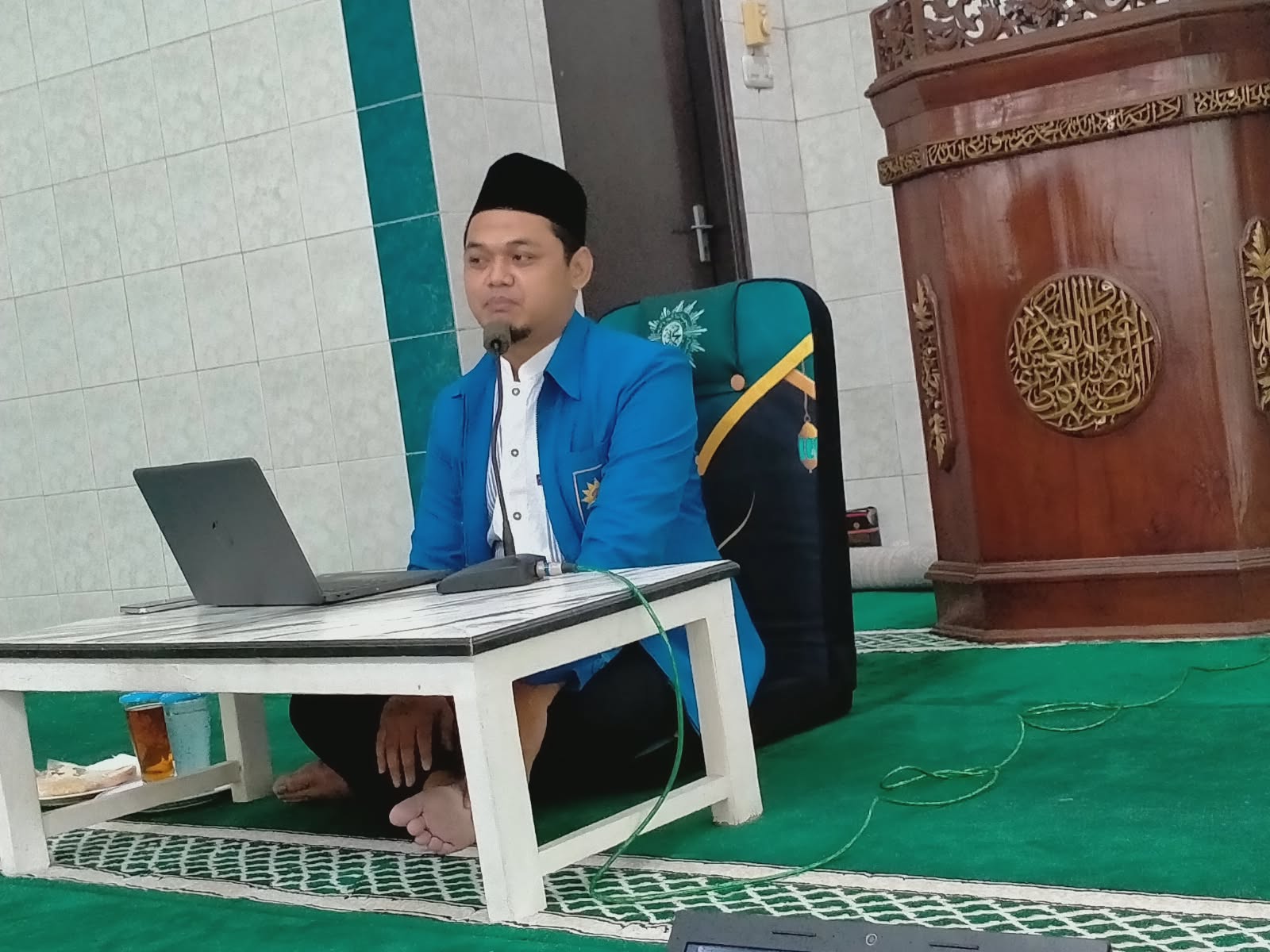 Foto seorang pria muda (Ustadz Amiril Edi Pranomo) mengenakan jas almamater berwarna biru cerah khas Muhammadiyah dengan kemeja putih dan peci hitam. Beliau duduk bersila secara lesehan di atas karpet masjid berwarna hijau, menghadap ke sebuah meja kayu kecil berwarna putih yang di atasnya terdapat sebuah laptop. Di sampingnya terlihat sebuah mimbar masjid yang terbuat dari kayu berwarna cokelat tua dengan ukiran kaligrafi Arab melingkar yang indah. Suasana ruangan tampak tenang dengan dinding keramik putih dan pilar berwarna hijau toska.