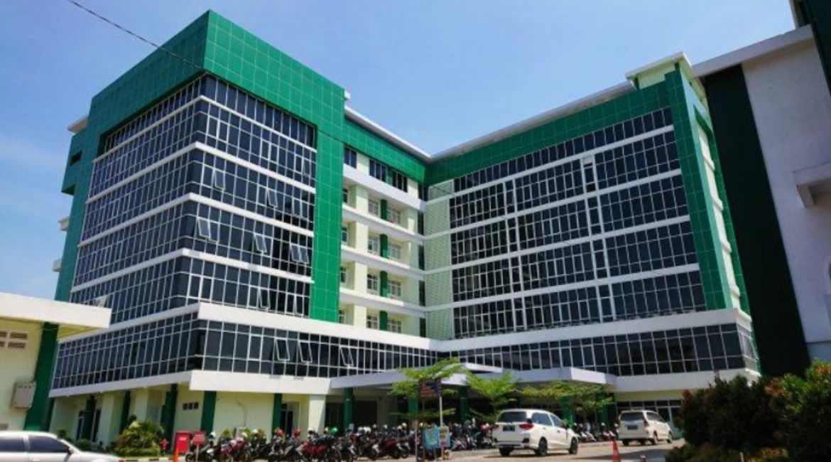 Foto sebuah gedung universitas modern yang megah dengan desain arsitektur kontemporer berbentuk huruf "L". Gedung ini memiliki tujuh lantai dengan dominasi fasad kaca gelap yang memberikan kesan profesional dan modern. Struktur bangunan dikelilingi oleh panel vertikal berwarna hijau cerah yang menjadi ciri khas identitas visual organisasi Muhammadiyah. Di area depan gedung, terdapat tempat parkir yang dipenuhi deretan sepeda motor dan beberapa mobil putih yang terparkir rapi. Langit biru yang cerah di latar belakang memberikan pencahayaan alami yang kuat pada seluruh kompleks bangunan.