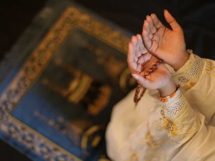 Foto close-up dari sudut pandang atas (high angle) yang memperlihatkan sepasang tangan seseorang yang sedang menadah berdoa. Orang tersebut mengenakan pakaian muslim berwarna putih dengan detail bordir kuning keemasan yang rumit di bagian lengan. Di antara kedua telapak tangannya, terselip sebuah tasbih kayu berwarna cokelat. Di latar belakang yang sedikit kabur (bokeh), tampak sebuah sajadah berwarna biru tua dengan ilustrasi gambar Kakbah dan menara Masjidil Haram yang detail. Pencahayaan foto terasa hangat dan fokus pada gerakan tangan yang melambangkan kekhusyukan.