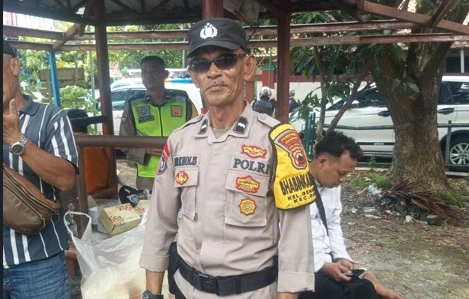 Aipda Nurkholis (nama di seragam NURHOLIS), petugas polisi paruh baya Bhabinkamtibmas Kelurahan Genuk, berdiri tegak mengenakan seragam dinas lengkap Polri, topi pet, kacamata hitam, dan ban lengan kuning, di luar ruangan di bawah gubug kayu dengan orang lain dan kendaraan di latar belakang.