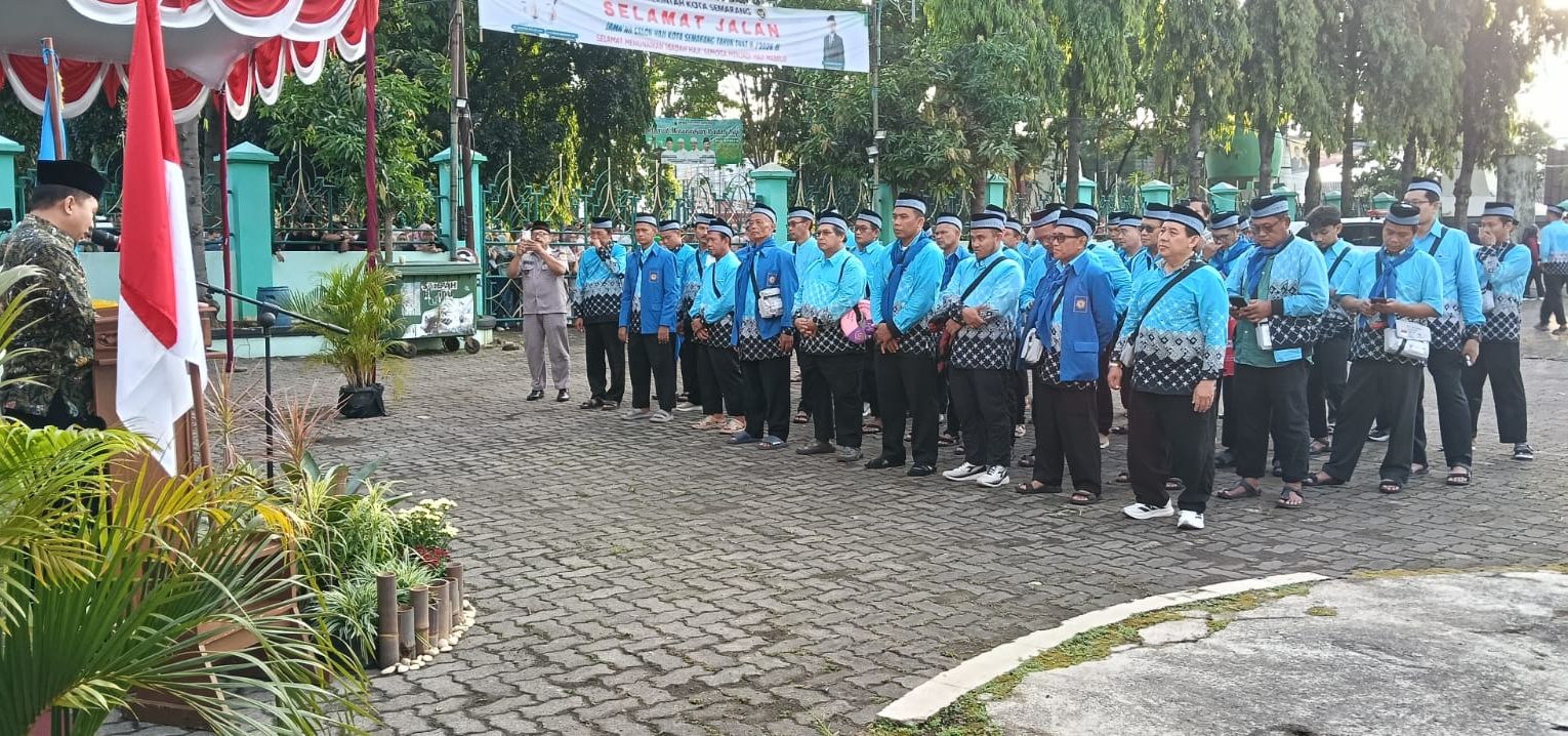 Foto menunjukkan barisan jemaah haji laki-laki yang mengenakan seragam batik haji nasional berwarna biru muda bermotif, dipadukan dengan jas biru tua dan peci hitam dengan aksen putih. Mereka berdiri dengan khidmat di sebuah lapangan berpaving. Di sebelah kiri, terdapat sebuah podium yang dihiasi bendera Merah Putih, di mana seorang pria berpakaian batik sedang memberikan sambutan. Di latar belakang, terlihat spanduk bertuliskan "Selamat Jalan" serta pepohonan hijau dan pagar besi hijau. Suasana tampak cerah dan tertib saat prosesi pelepasan berlangsung.