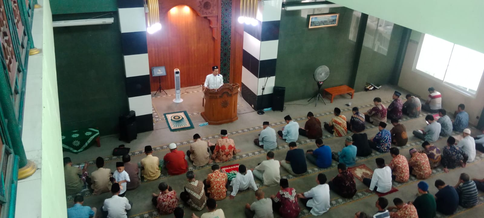 Foto yang diambil dari sudut pandang tinggi (high angle) menunjukkan suasana bagian dalam sebuah masjid yang didominasi warna hijau dan putih. Di bagian depan, seorang khatib mengenakan baju koko putih dan peci hitam berdiri di atas mimbar kayu berwarna cokelat yang memiliki ukiran matahari khas Muhammadiyah. Di hadapannya, puluhan jamaah pria duduk rapi di atas karpet hijau, mengenakan berbagai jenis pakaian seperti kemeja batik dan baju koko. Ruangan masjid memiliki pilar-pilar besar dengan motif garis hitam-putih, serta pencahayaan yang terang dari lampu gantung di area mihrab.