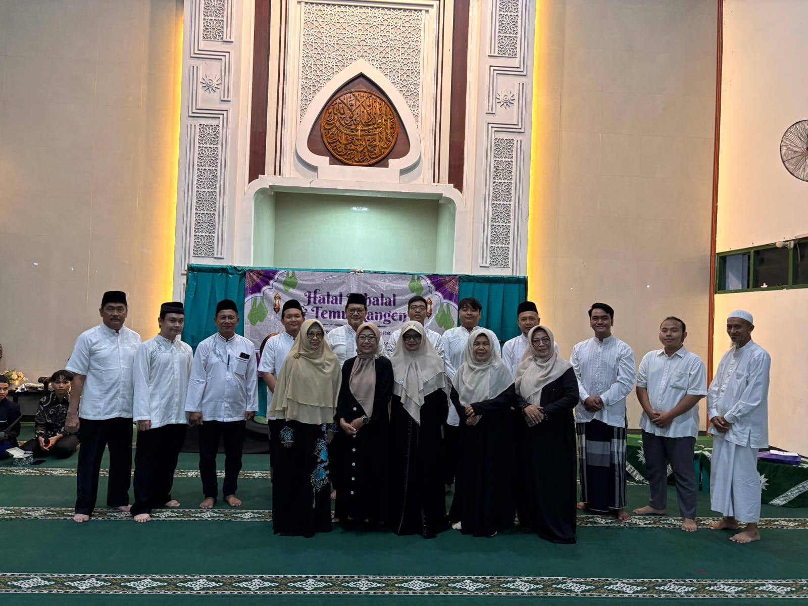 Foto bersama sekelompok pria dan wanita di dalam ruang utama sebuah masjid yang memiliki langit-langit tinggi dan dinding berwarna krem netral. Di latar belakang, terdapat mihrab yang megah dengan ukiran kaligrafi Arab berbentuk lingkaran di bagian tengah dan pola geometris yang rumit di sekelilingnya, diapit oleh dua pilar dengan pencahayaan lampu kuning yang hangat. Para pria mengenakan baju koko putih dan peci hitam, sementara para wanita mengenakan abaya hitam dengan kerudung berwarna krem atau cokelat muda. Di depan mihrab, terdapat spanduk bertuliskan "Halal Bihalal". Lantai masjid ditutupi karpet berwarna hijau tua dengan pola sajadah.