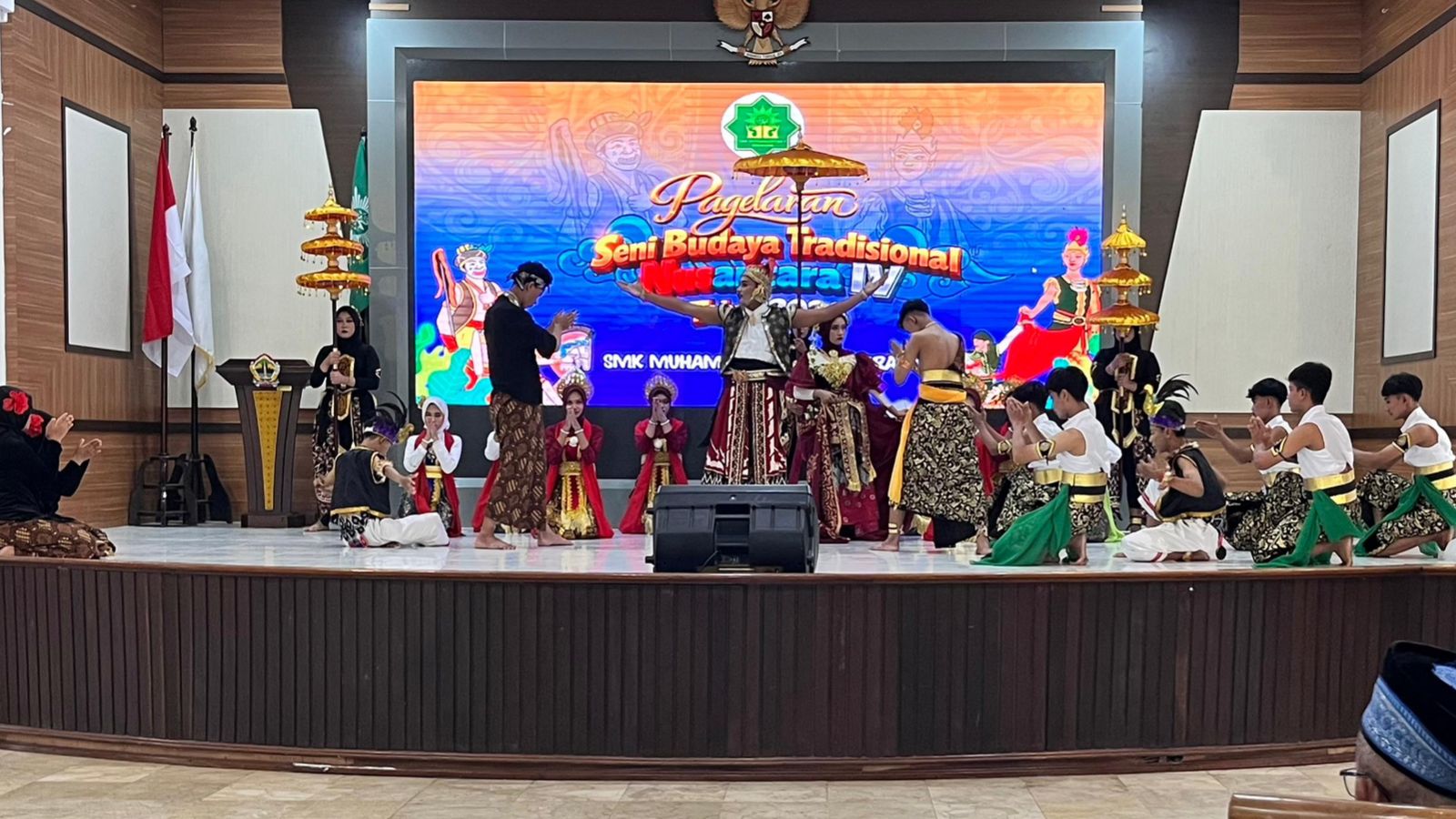 Foto panggung utama di dalam sebuah gedung yang menampilkan pertunjukan seni tradisional oleh sekelompok siswa pria dan wanita. Di bagian tengah panggung, seorang siswa pria berdiri gagah mengenakan kostum adat Jawa lengkap dengan kain batik, rompi, dan aksesori kepala, dikelilingi oleh penari lain yang duduk bersimpuh dalam posisi hormat. Di latar belakang, terdapat layar LED besar bertuliskan "Pagelaran Seni Budaya Tradisional Nusantara IV SMK Muhammadiyah 1 Semarang". Panggung dihiasi dengan payung tradisional berwarna kuning keemasan di sisi kiri dan kanan, serta tanaman hias di bagian depan panggung. Suasana ruangan tampak formal dan megah dengan langit-langit berdesain modern.