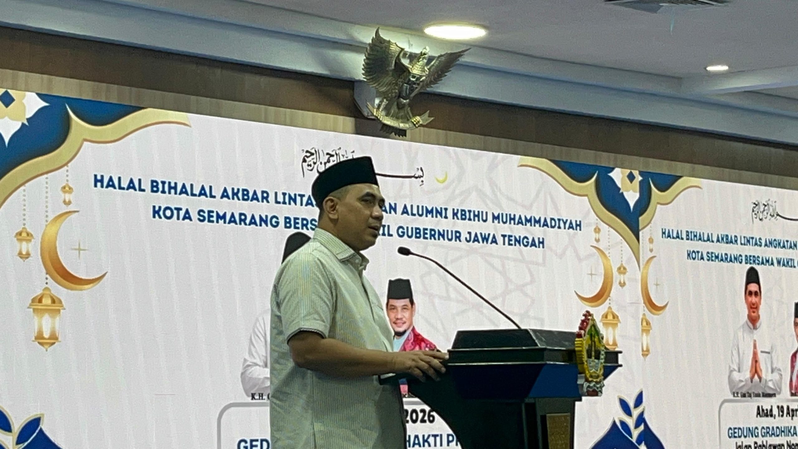 Foto Wakil Gubernur Jawa Tengah, K.H. Taj Yasin Maimoen, yang sedang memberikan sambutan dari balik podium hitam dengan logo emas di bagian depan. Beliau mengenakan kemeja berwarna krem muda bermotif garis halus dan peci hitam. Di latar belakang, terdapat layar LED besar yang menampilkan teks "HALAL BIHALAL AKBAR LINTAS ANGKATAN ALUMNI KBIHU MUHAMMADIYAH KOTA SEMARANG BERSAMA WAKIL GUBERNUR JAWA TENGAH" dengan dekorasi nuansa Islami berupa gambar bulan sabit dan lampu lampion. Di bagian atas tengah layar, terdapat kaligrafi Arab. Suasana ruangan tampak formal dengan pencahayaan lampu langit-langit yang terang.