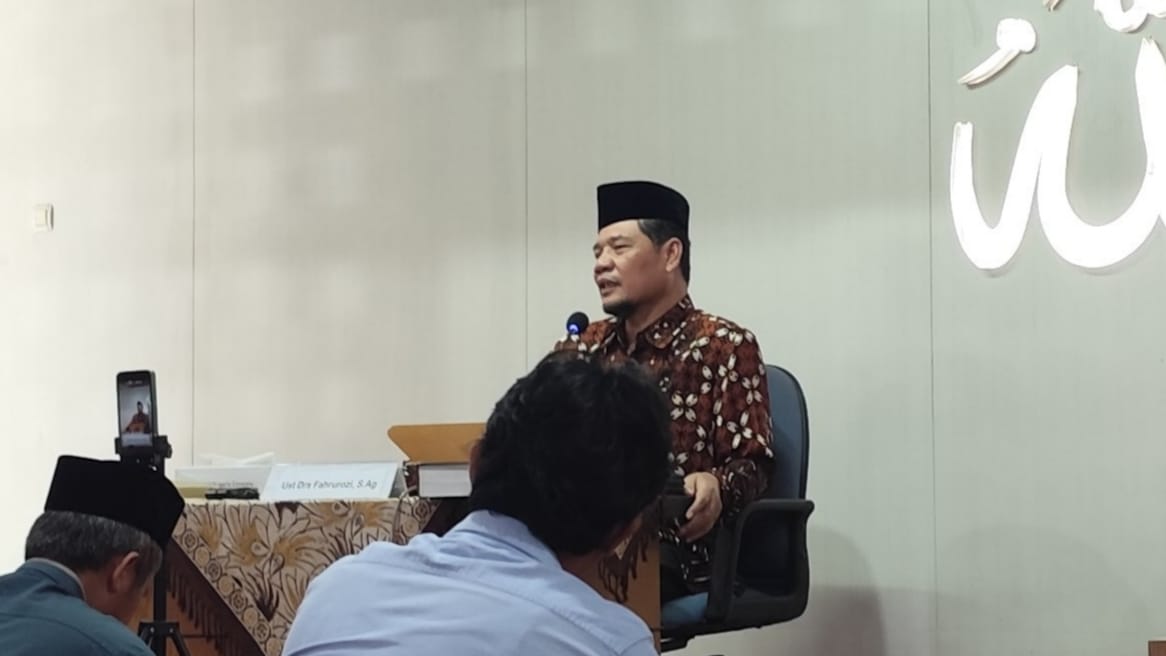 Foto yang menunjukkan Ust. Dr. Fachrur Rozi, M.Ag. sedang memberikan tausiyah dalam posisi duduk di sebuah kursi kantor berwarna hitam. Beliau mengenakan kemeja batik bermotif cokelat tua dan peci hitam, berbicara melalui mikrofon di sebuah ruangan yang memiliki dinding putih bersih. Di sebelah kanan beliau, terdapat dekorasi dinding berupa kaligrafi Arab menyala yang membentuk tulisan "Allah". Di depan beliau, terlihat bagian belakang kepala seorang jemaah yang mengenakan kemeja biru, serta sebuah meja kayu yang dilapisi taplak batik cokelat dengan tumpukan buku dan perangkat ponsel yang diletakkan di atas tripod untuk mendokumentasikan kajian.
