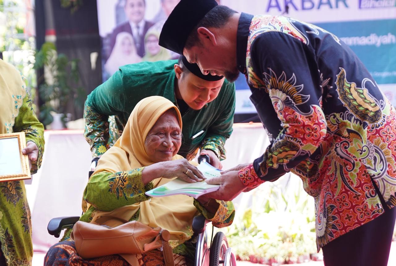Foto candid yang penuh haru memperlihatkan seorang wanita lansia (Ibu Hj. Siti Murtini Mashuri) mengenakan jilbab kuning dan pakaian hijau bermotif bunga, sedang duduk di kursi roda. Beliau tampak tersenyum lembut saat menerima sebuah buku atau dokumen dari seorang pria muda yang mengenakan kemeja batik bermotif burung phoenix berwarna-warni. Di belakang beliau, seorang pria lain berbaju hijau membantu memegang sandaran kursi roda dengan penuh perhatian. Di sisi kiri foto, tampak tangan orang lain yang memegang bingkai penghargaan berwarna emas. Latar belakang foto memperlihatkan suasana acara luar ruangan yang cerah dengan spanduk acara "Hari BerMuhammadiyah".