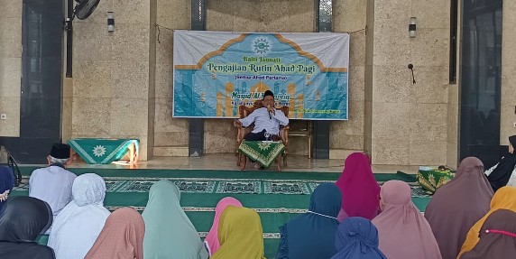 Seorang penceramah pria duduk di panggung menyampaikan tausiyah Halal Bi Halal Majelis Ta’lim Baiti Jannati di Masjid Al Muhajirin Karangayu Semarang. Di belakangnya terdapat spanduk acara. Jemaah, pria dan wanita berhijab, duduk di karpet di depan.