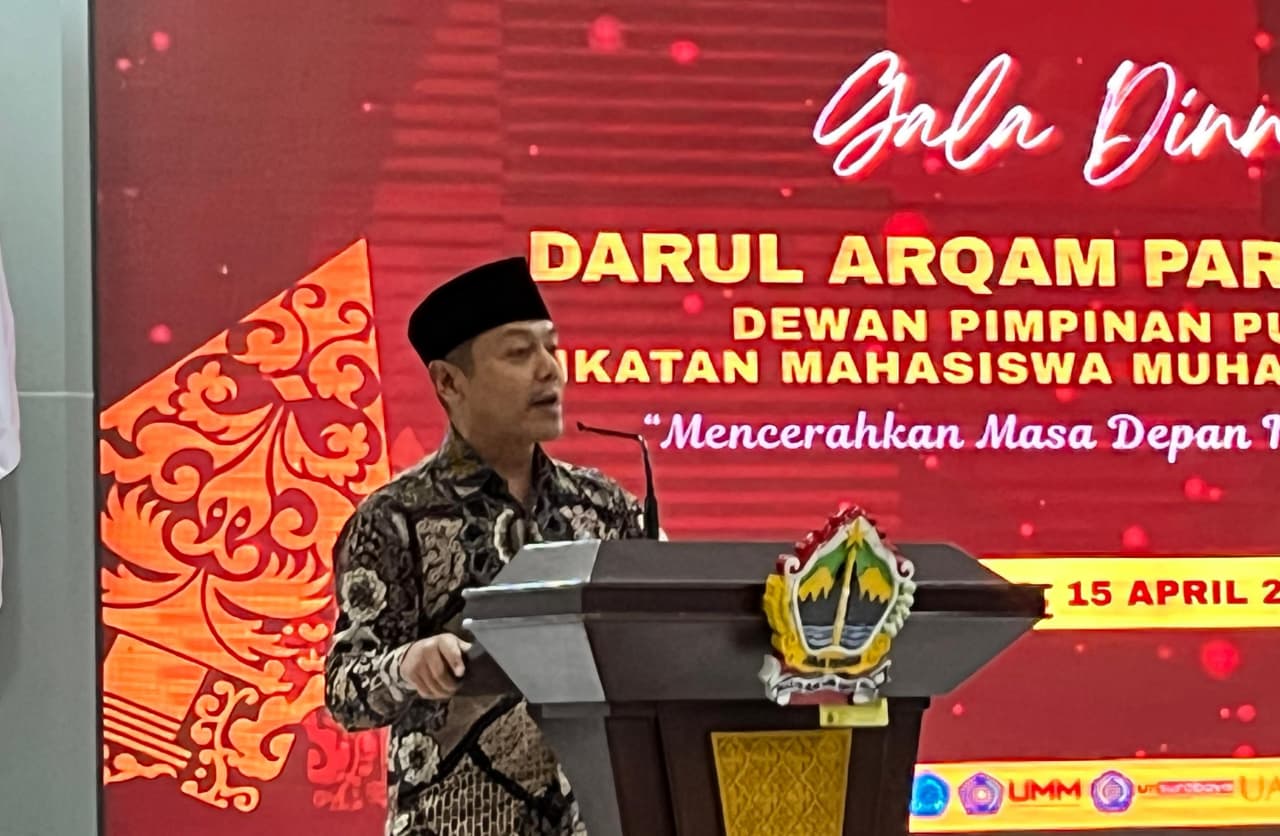 Foto close-up Wakil Menteri Pendidikan Dasar dan Menengah (Wamen Dikdasmen), Dr. Fajar Riza Ul Haq, M.A., yang sedang memberikan sambutan di balik podium kayu dengan logo universitas. Beliau mengenakan batik motif parang berwarna gelap dan peci hitam. Di latar belakang, terdapat layar LED besar berwarna merah dengan teks putih yang memuat tulisan "Gala Dinner DARUL ARQAM PARIPURNA" dan "Mencerahkan Masa Depan Indonesia". Suasana pencahayaan fokus pada sosok pembicara yang sedang memaparkan materi.