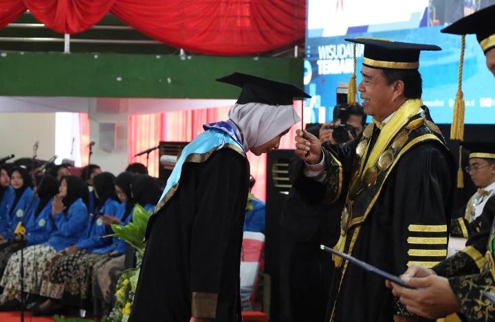 Foto suasana wisuda di dalam gedung pertemuan yang besar. Terlihat seorang wisudawan wanita mengenakan jilbab putih, toga hitam dengan samir (kalung wisuda) berwarna biru muda dan emas, sedang menundukkan kepala dengan hormat di depan panggung. Di hadapannya, Rektor Unimus, Prof. Dr. Masrukhi, M.Pd., yang mengenakan toga rektor hitam dengan kalung medali emas besar dan topi wisuda emas, sedang memindahkan kucir topi wisudawan tersebut dari kiri ke kanan. Di latar belakang, tampak layar LED besar menampilkan tulisan "WISUDAWAN TERBAIK". Puluhan wisudawan lain mengenakan jaket almamater biru duduk lesehan di barisan kursi penonton di bawah dekorasi kain merah. Seorang fotografer terlihat sedang mengabadikan momen tersebut di panggung.